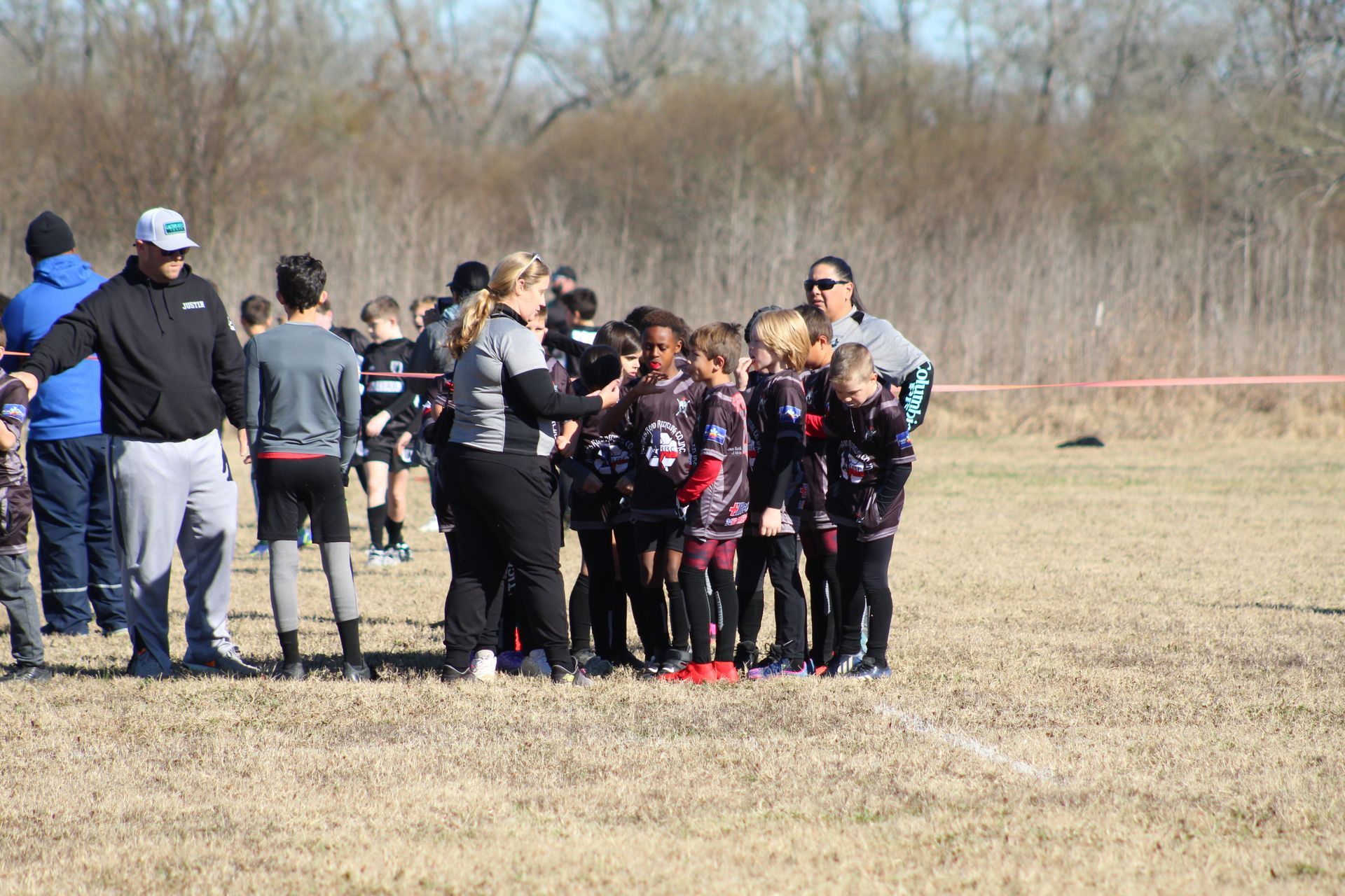A youth soccer team huddles with coaches on a dry, grassy field on a sunny day.