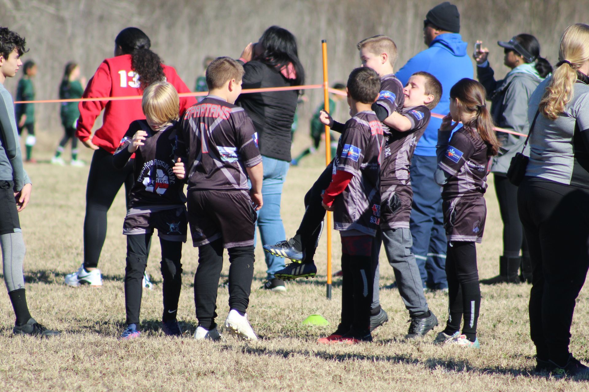 Youth soccer players in black and purple jerseys huddle together on a grassy field, with spectators in the background.