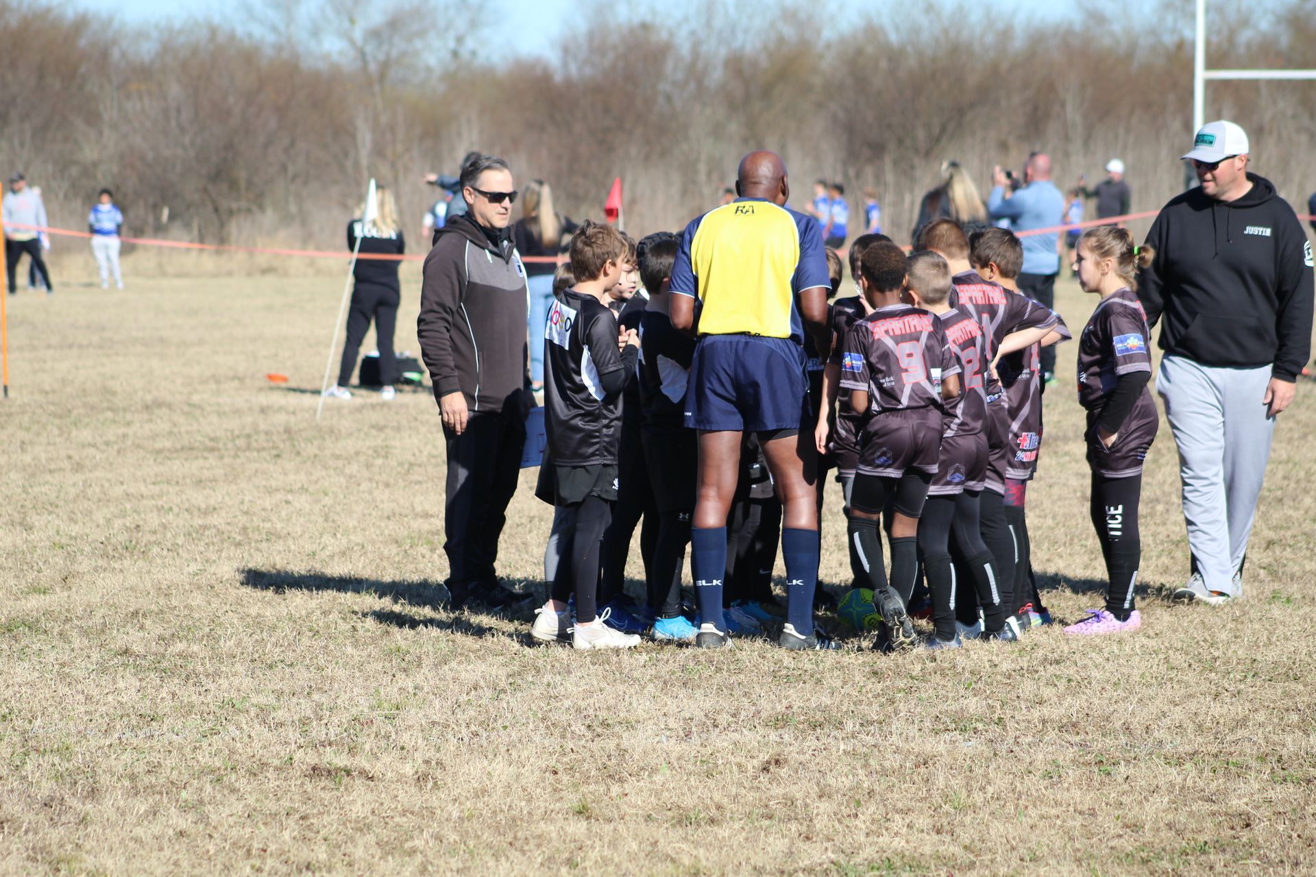 Rugby team huddles with referee on a grassy field; coaches watch nearby.