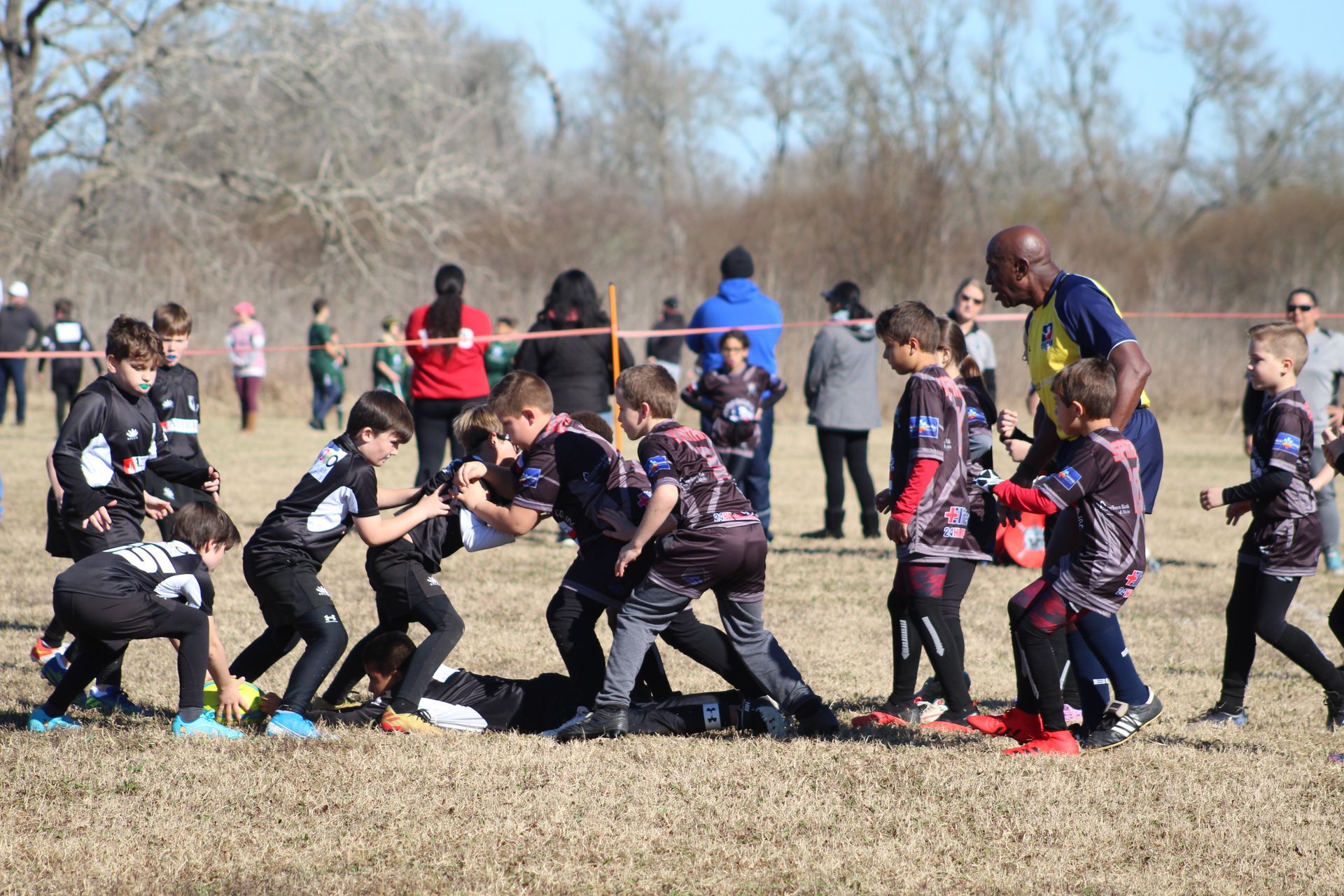 Youth rugby players in black jerseys tackle on a grassy field; coach watches.
