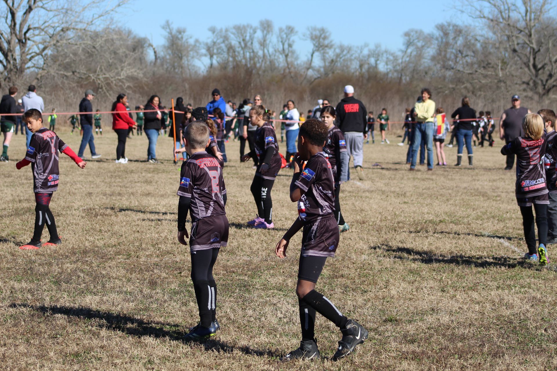 Children in black and pink athletic attire on a grassy field, spectators in the background.