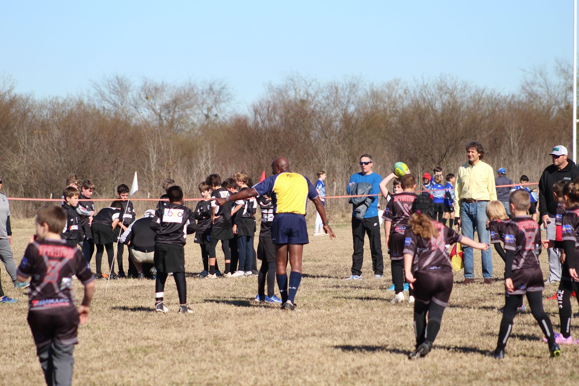 Rugby players in black and pink uniforms huddle around a referee on a dry field under a clear sky.