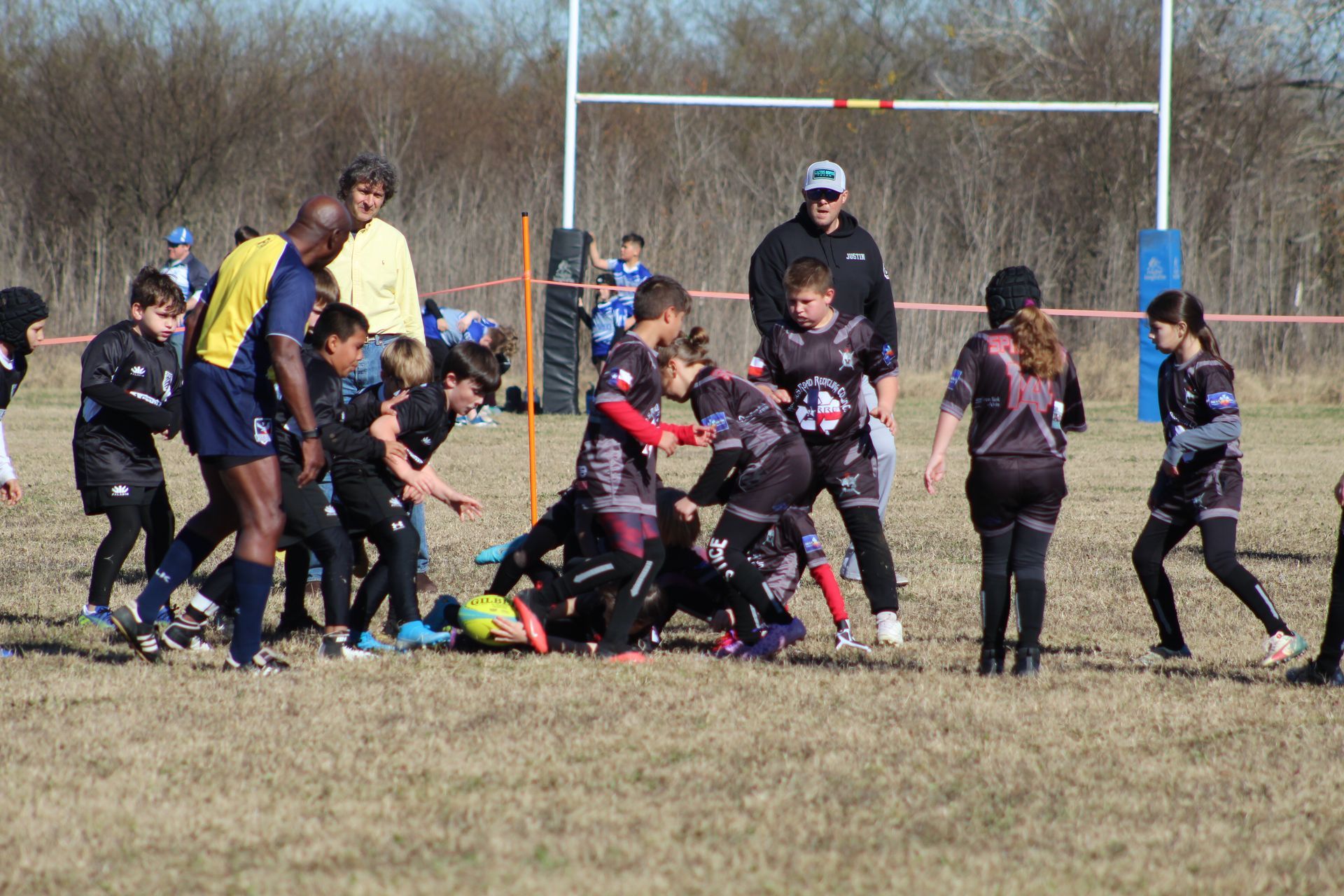 Children in rugby uniforms on a field, scrumming around a ball; coaches and a goalpost visible.