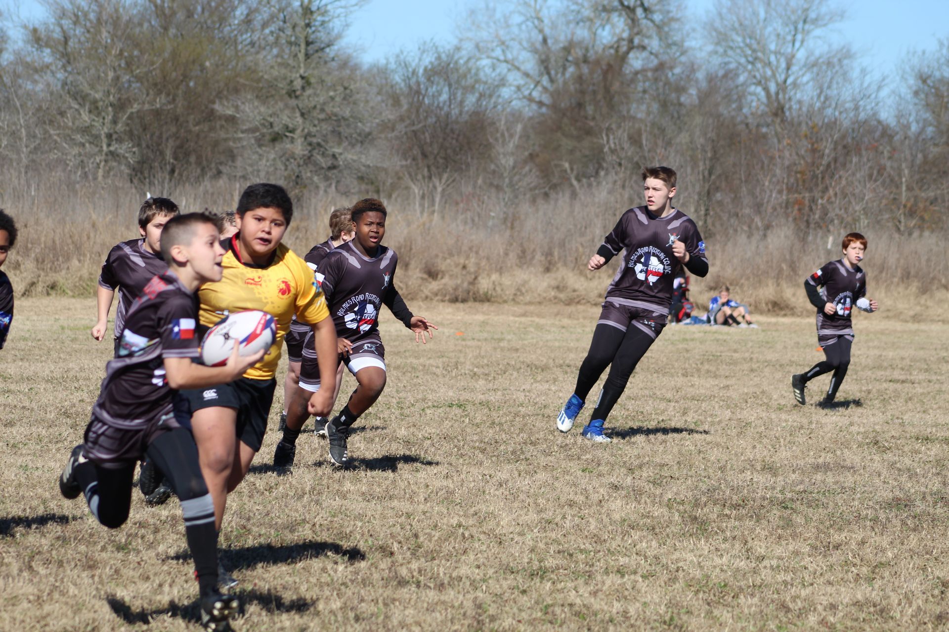Rugby players running on a field, some in black jerseys with white and blue details, one in a yellow jersey