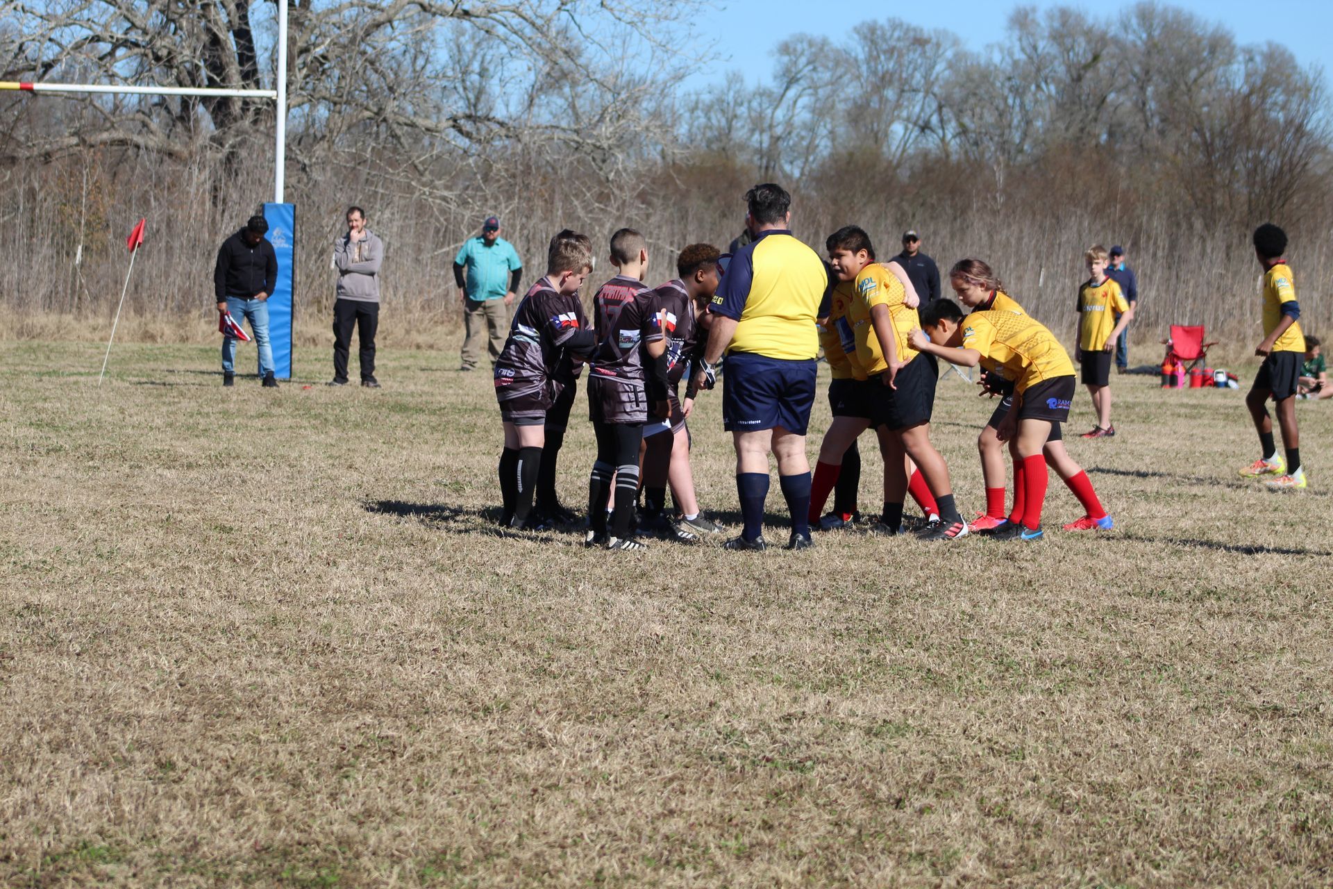 Rugby game in progress. Players in yellow and black jerseys huddle near the center field; spectators watch.
