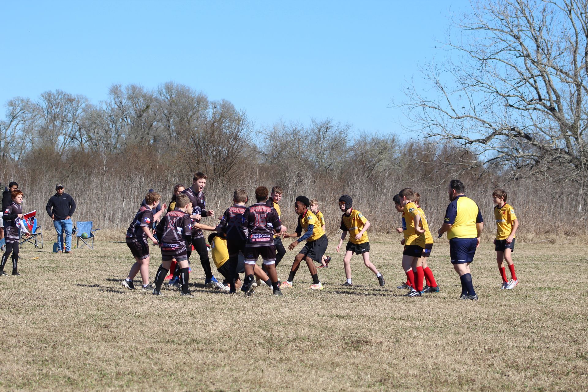 Rugby players in black and gold jerseys engaged in a scrum on a field under a blue sky.