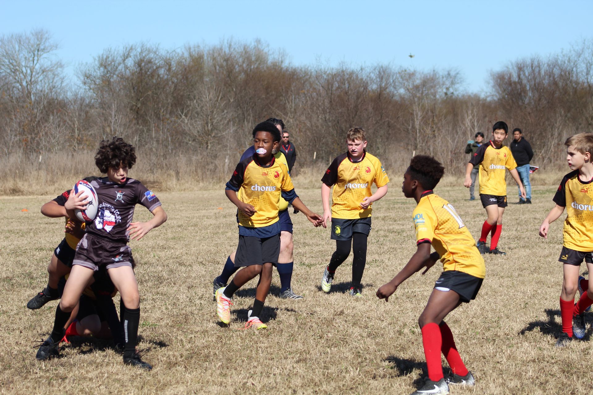 Rugby players in yellow and black uniforms on a field. One player with a ball is tackled.