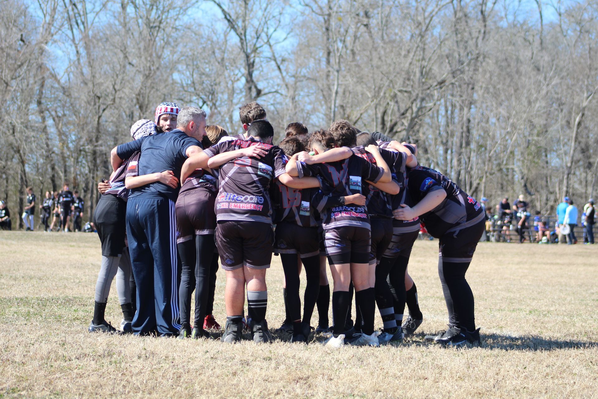 Rugby team in black uniforms huddling on a grassy field, arms around each other.