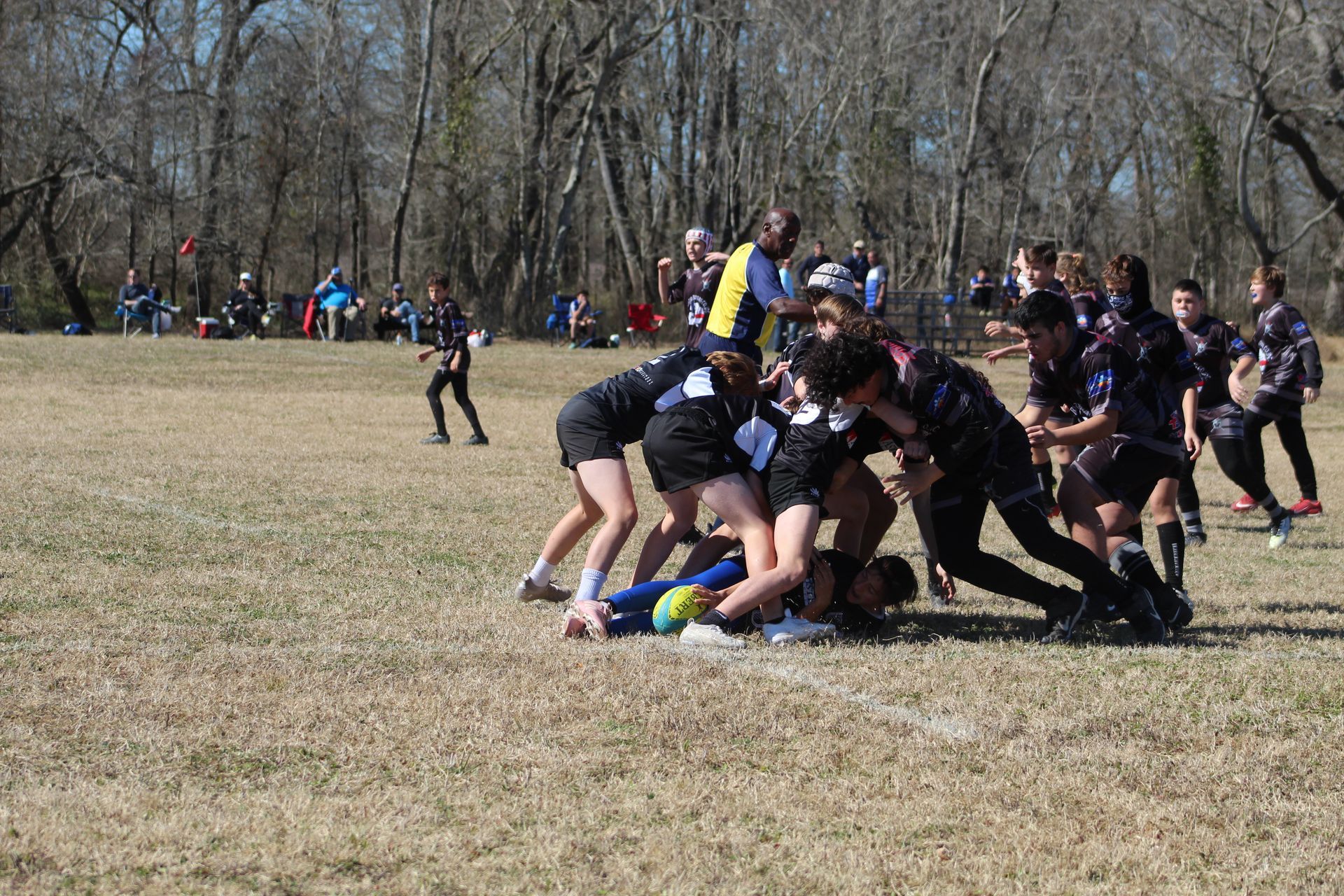 Rugby players in black and white uniforms engaged in a scrum on a grassy field; spectators in the background.