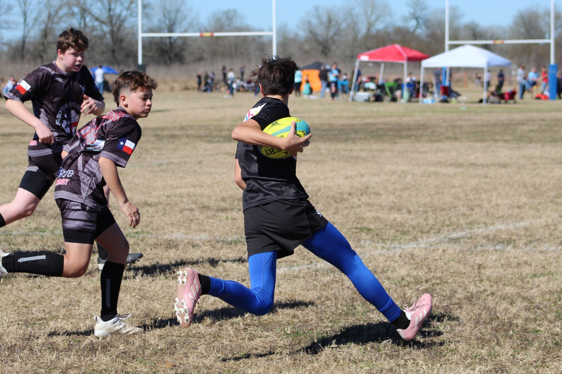 Rugby player runs with ball on field, other players nearby. Sunny day, open field, goal posts visible.