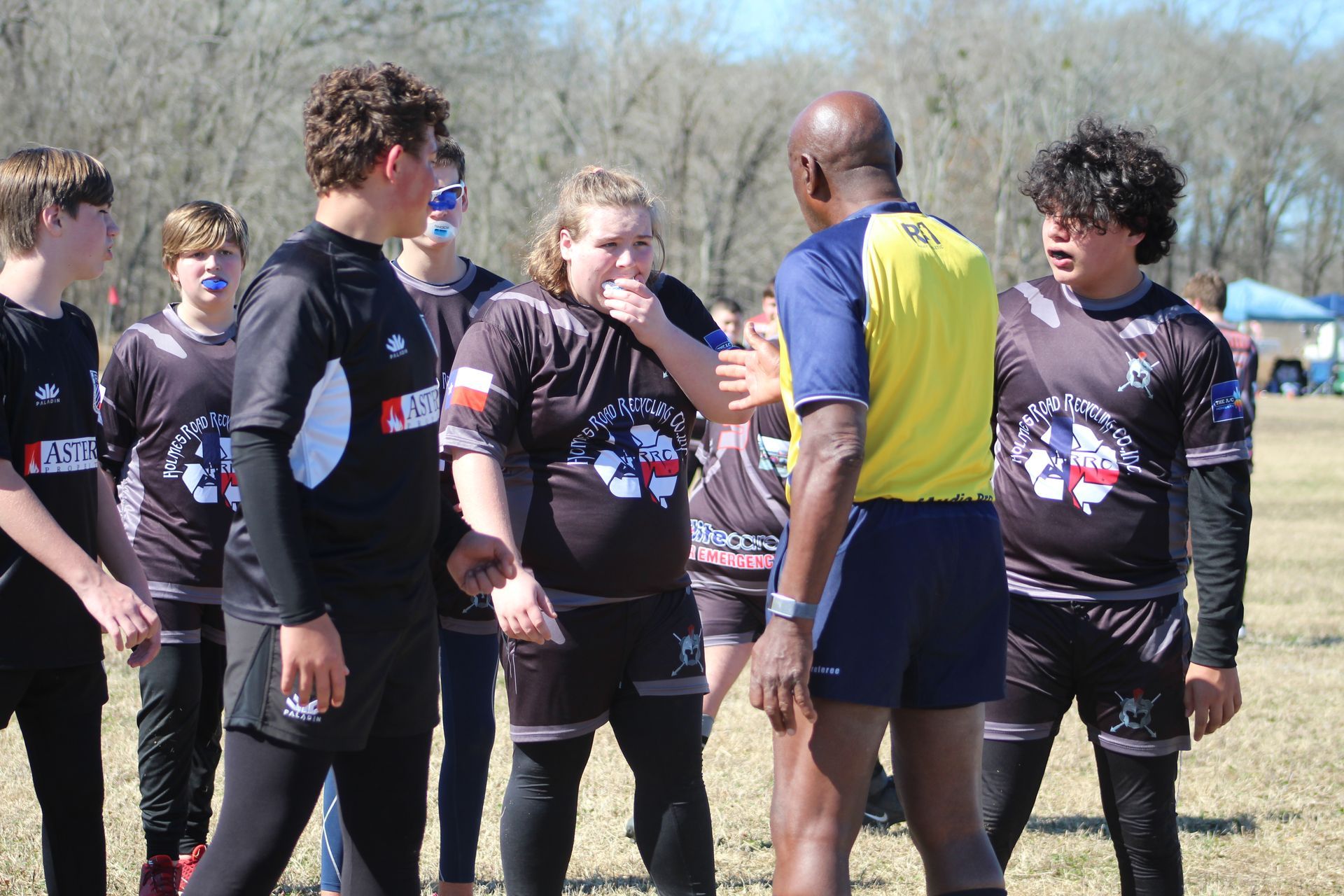 Rugby players in black jerseys huddle near a referee, outdoors on a sunny day.