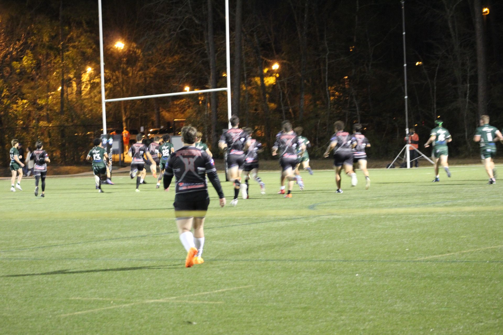 Rugby players in black and pink jerseys running on a green field towards goalposts at night.