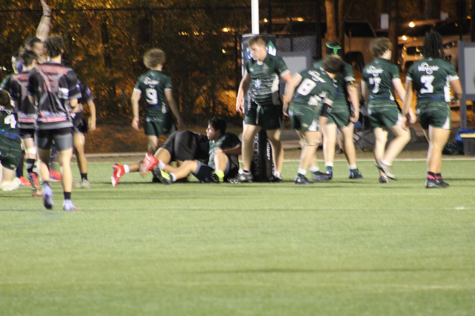 Rugby players in green and black uniforms on a field at night. One player tackled. Spectators watch.
