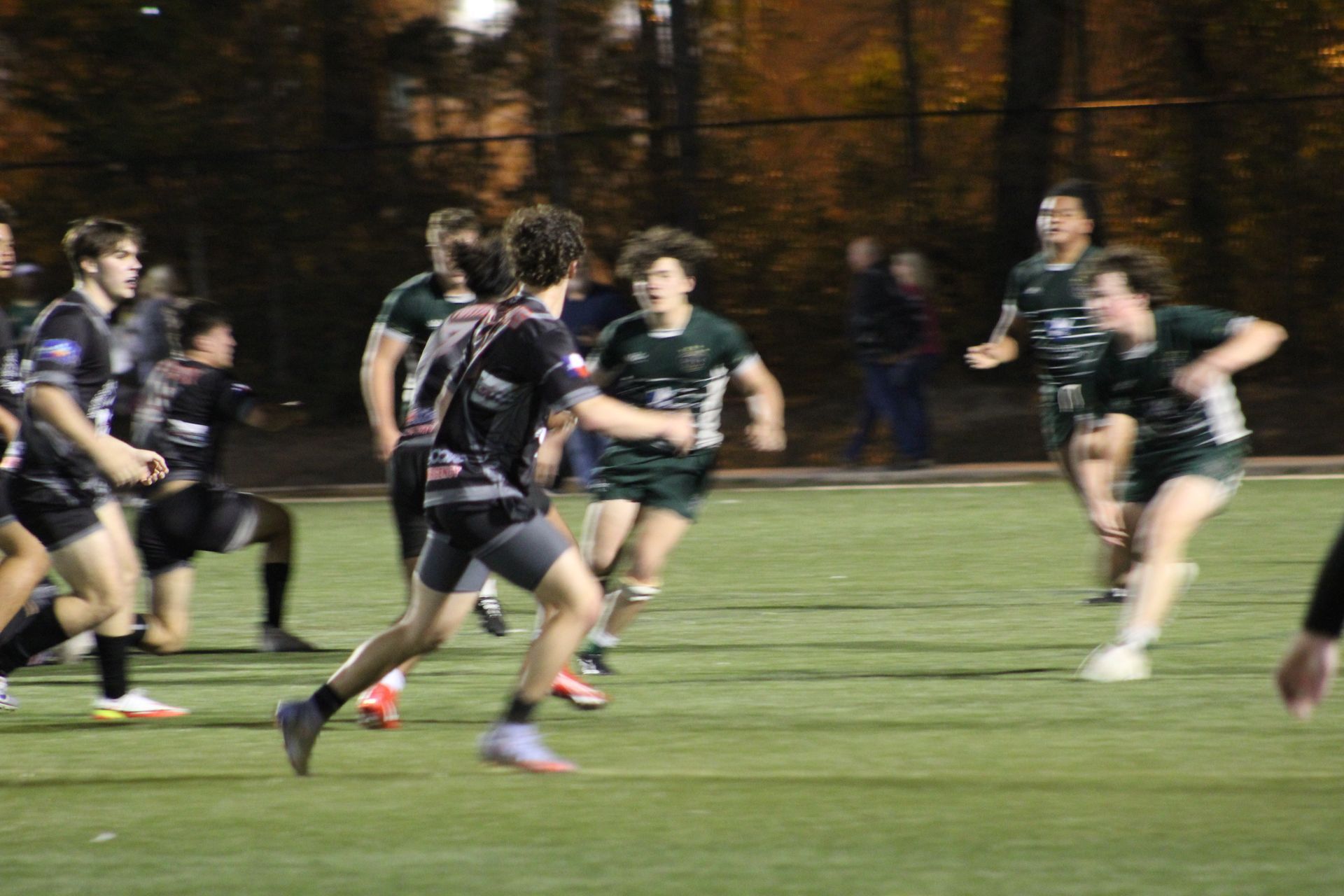 Rugby players on green field. One player in black runs forward, others in green chase. Night game.