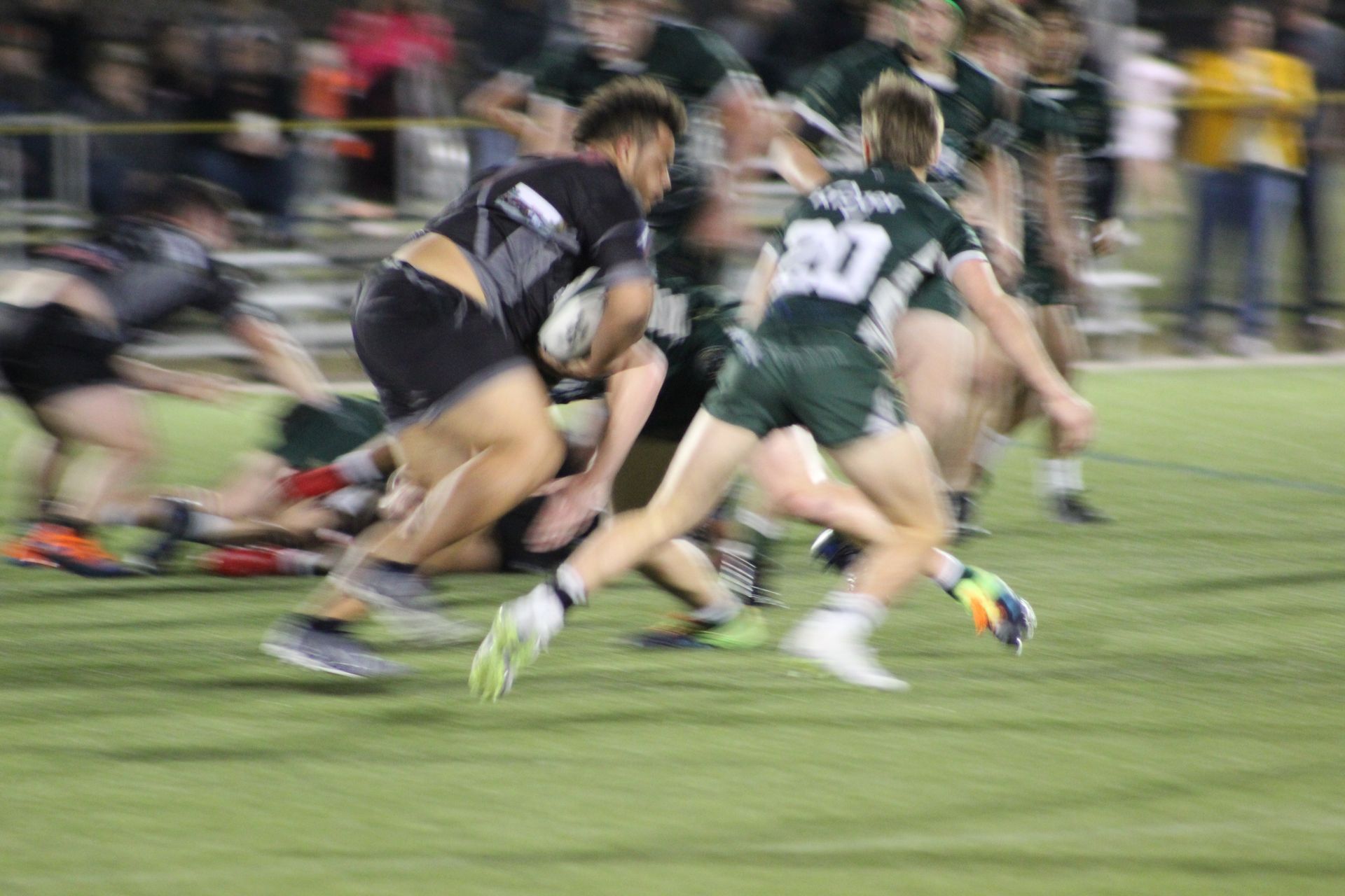 Rugby players in action; one with the ball being tackled; green and black uniforms on a field at night.
