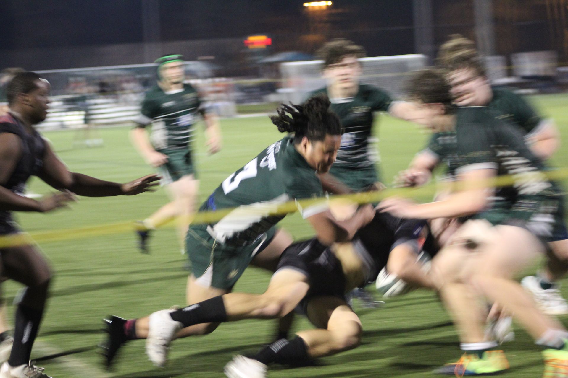 Rugby players in green and black uniforms engaged in a tackle on a green field at night.