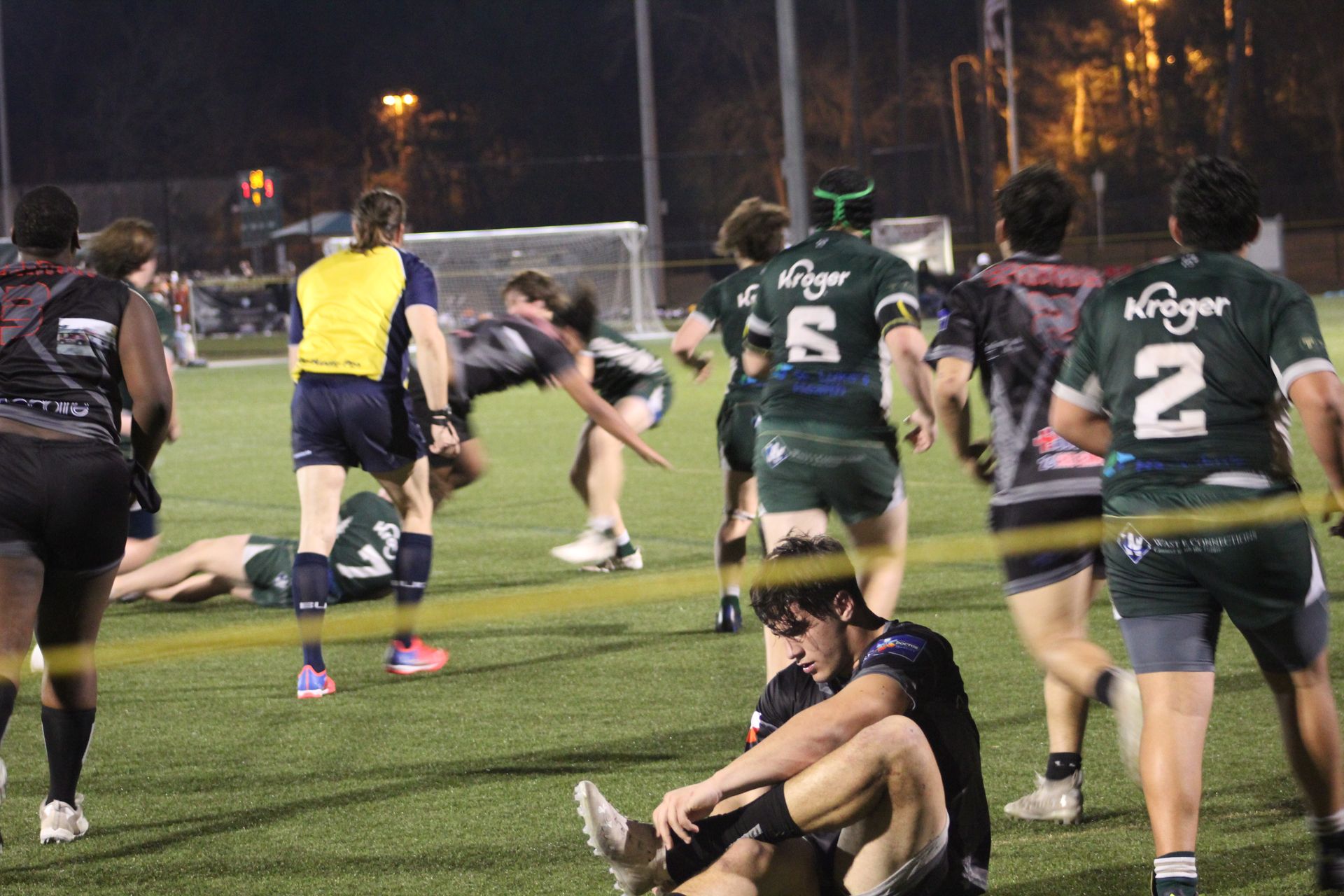 Rugby players in green and black uniforms on a field at night. One player sits on the ground, others run.