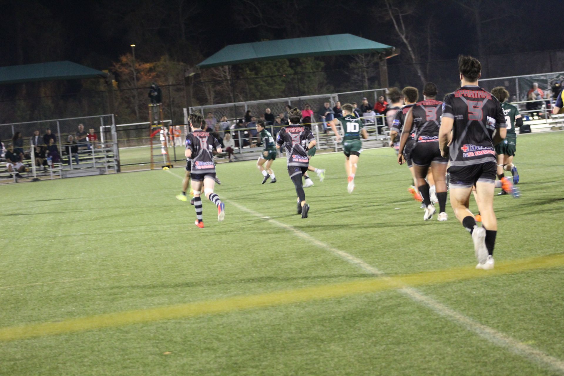 Rugby players in black and gray jerseys running on a green field at night, spectators in the background.