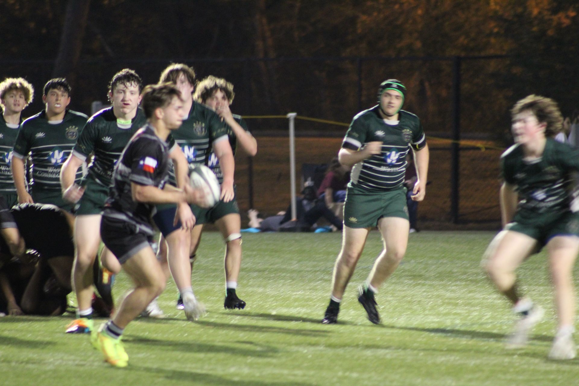 Rugby players on a field at night; one in black passes the ball, others in green watch and run.