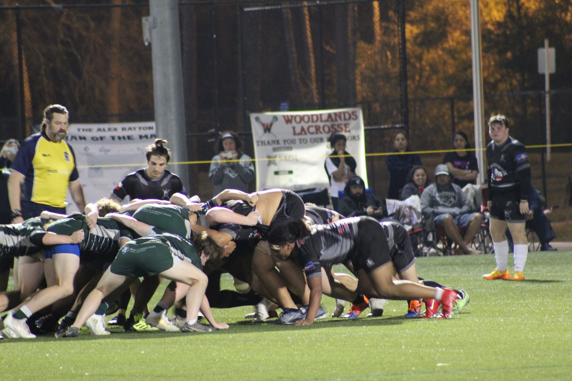 Rugby scrum on a green field at night. Players in uniforms, some watching, others are in a tight huddle.