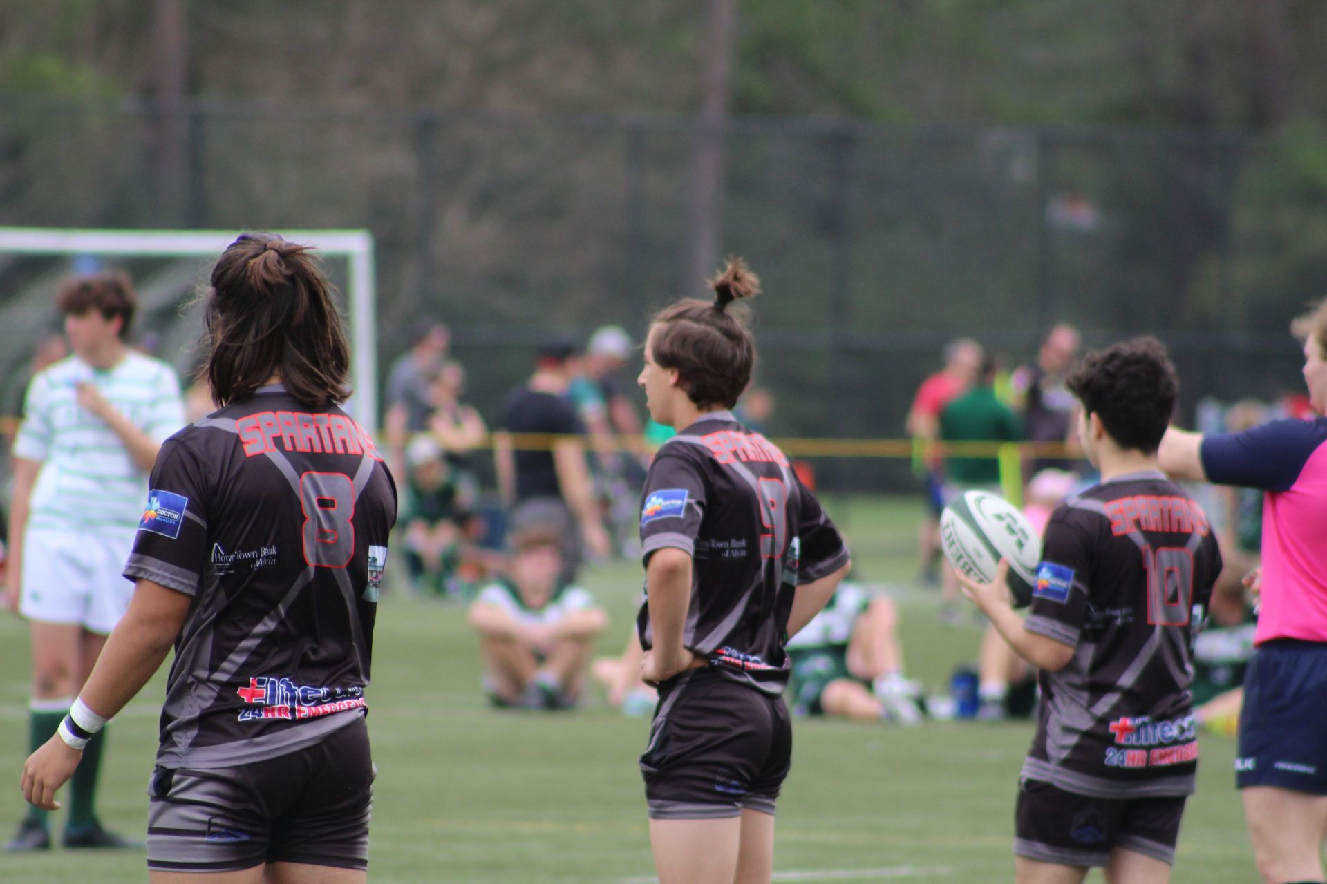 Rugby players in black jerseys on a green field. One holds the ball, others watch. Spectators in the background.
