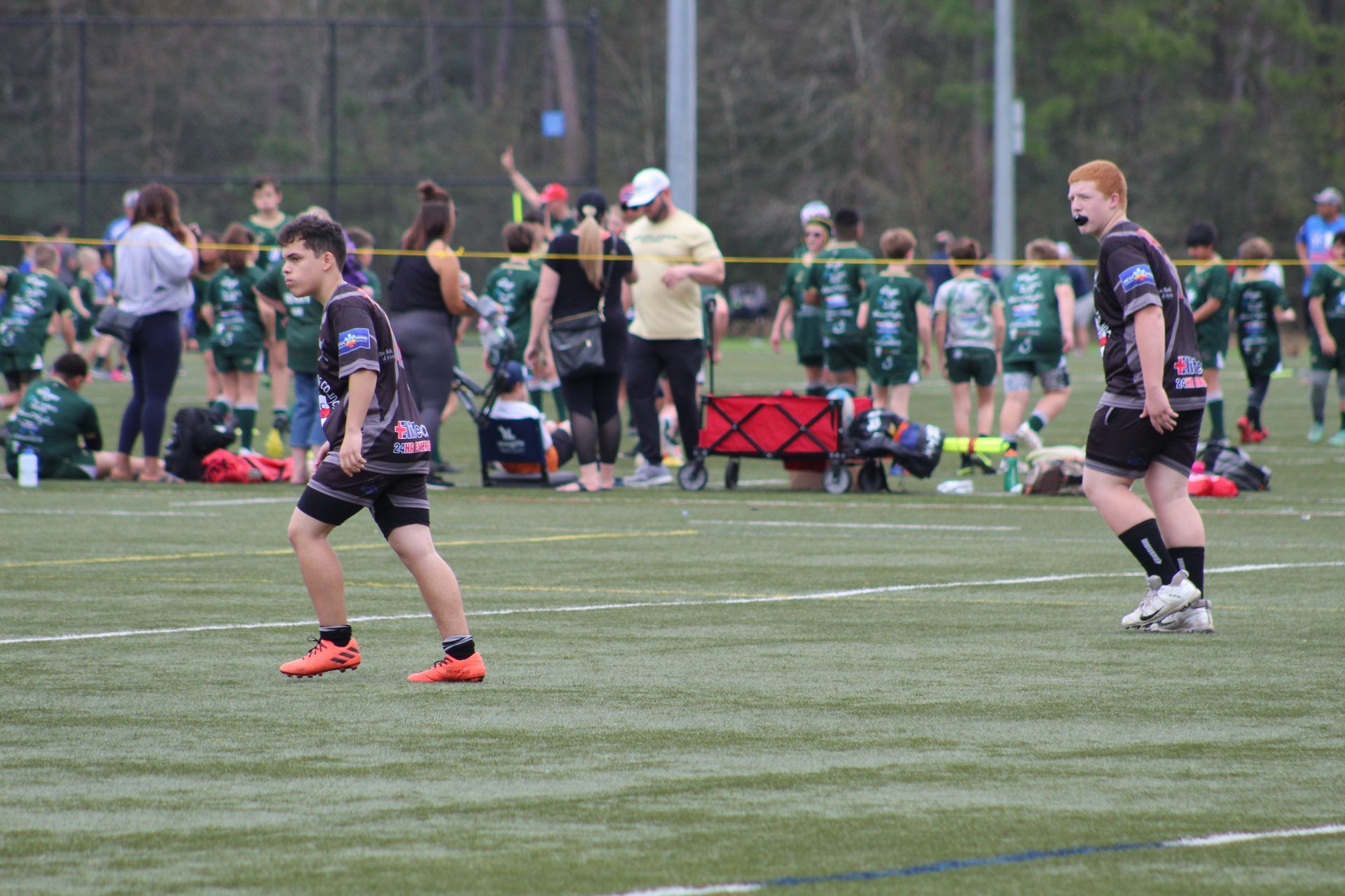 Two rugby players in black jerseys on a green field. Spectators and a red wagon are in the background.