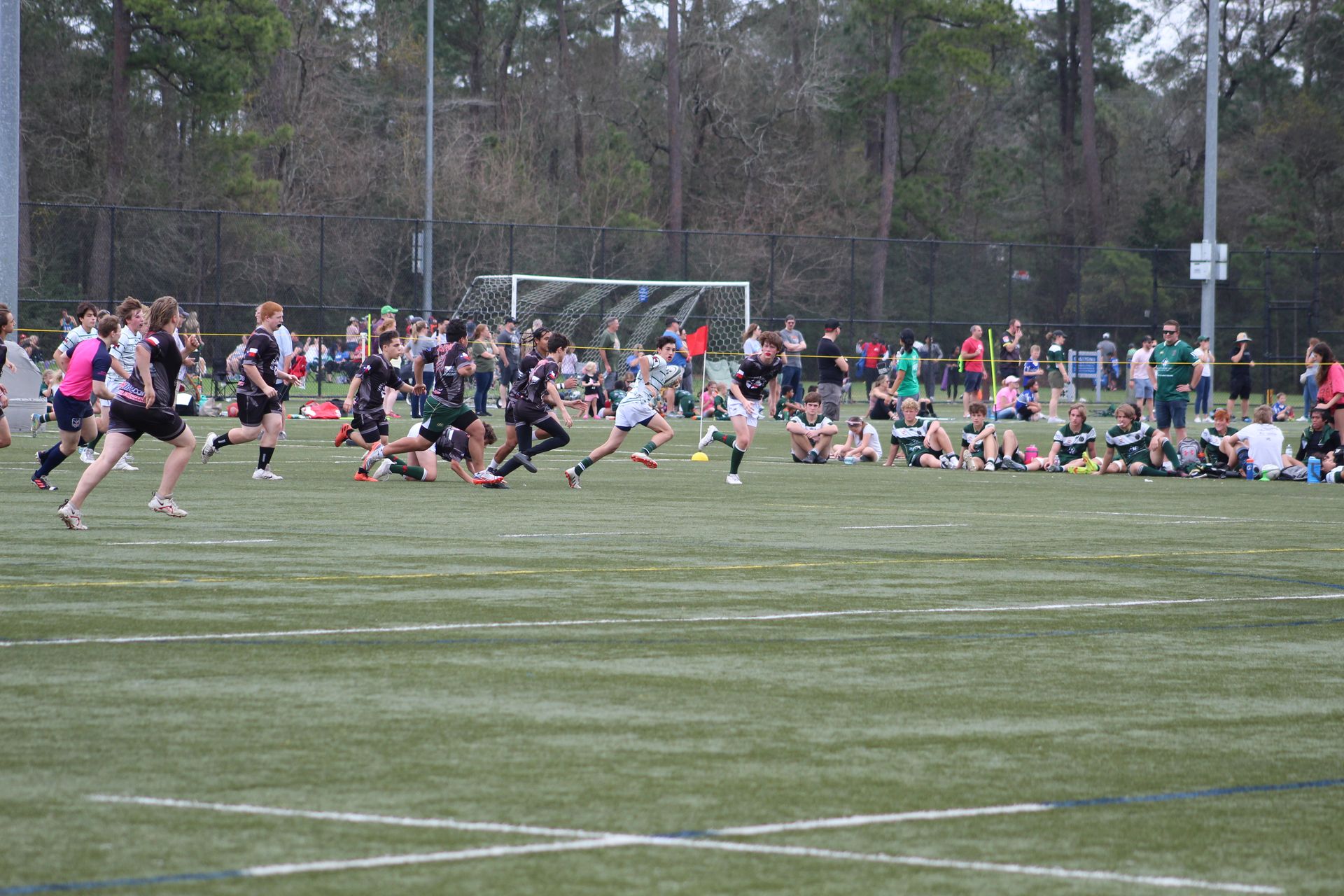 Rugby match on a green field; players in dark and light uniforms running and tackling. Spectators watch.