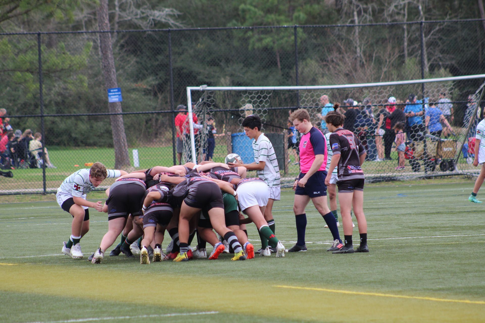Rugby players in a scrum, on a green field with spectators and a goal post in the background.
