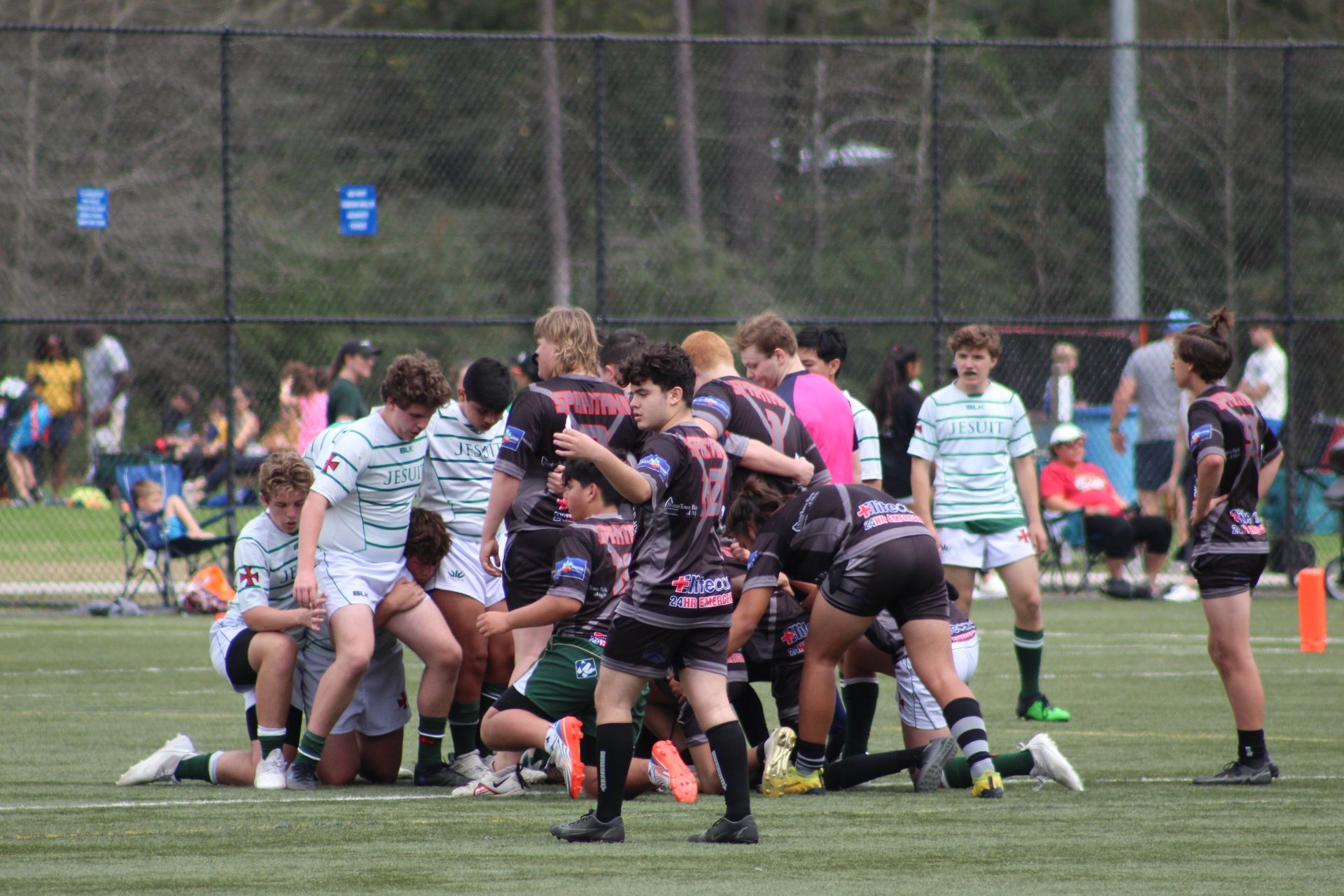 Rugby players in green and black jerseys huddle on a green field. Others watch in the background.