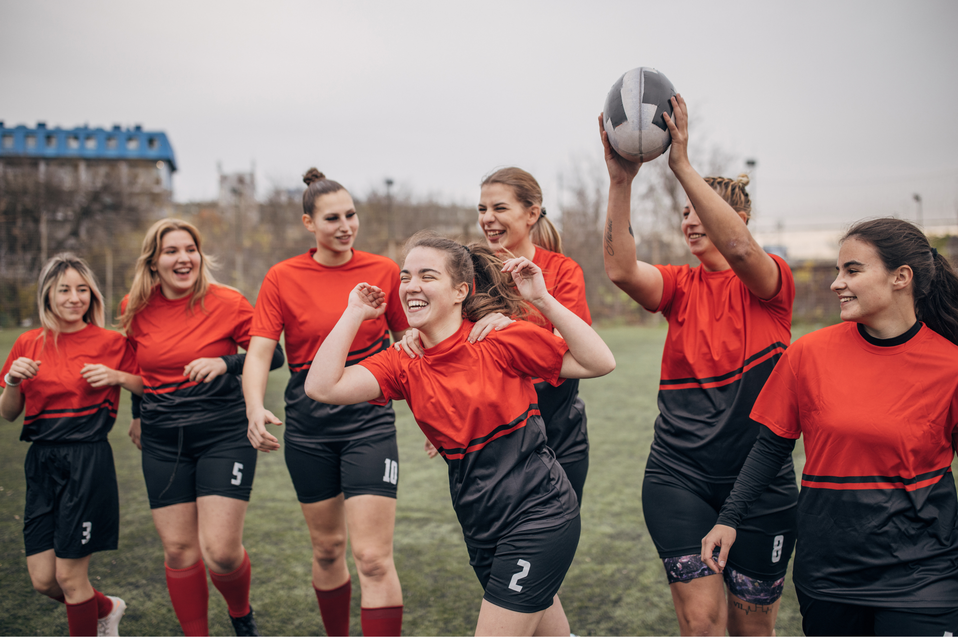 Women's rugby team celebrating a win on a green field. They wear red and black uniforms, holding a ball, and cheering.