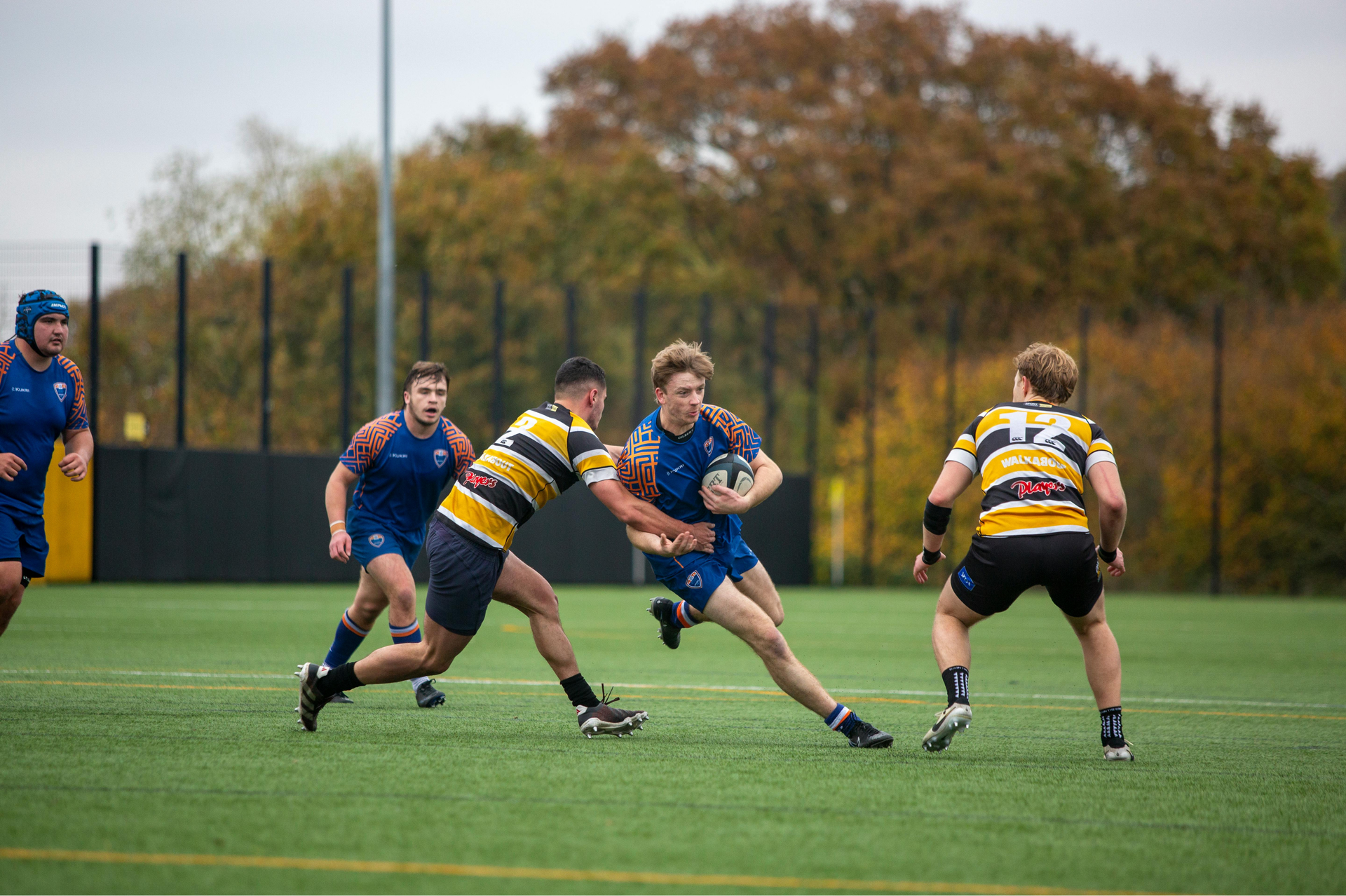 Rugby players in action; blue jersey runner tackled by striped jersey defender, green field, autumn trees.