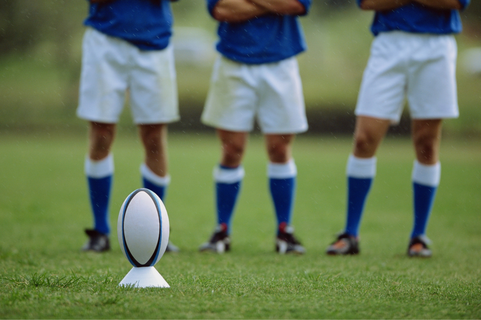 Rugby ball on a tee with three players in blue and white uniforms standing behind.