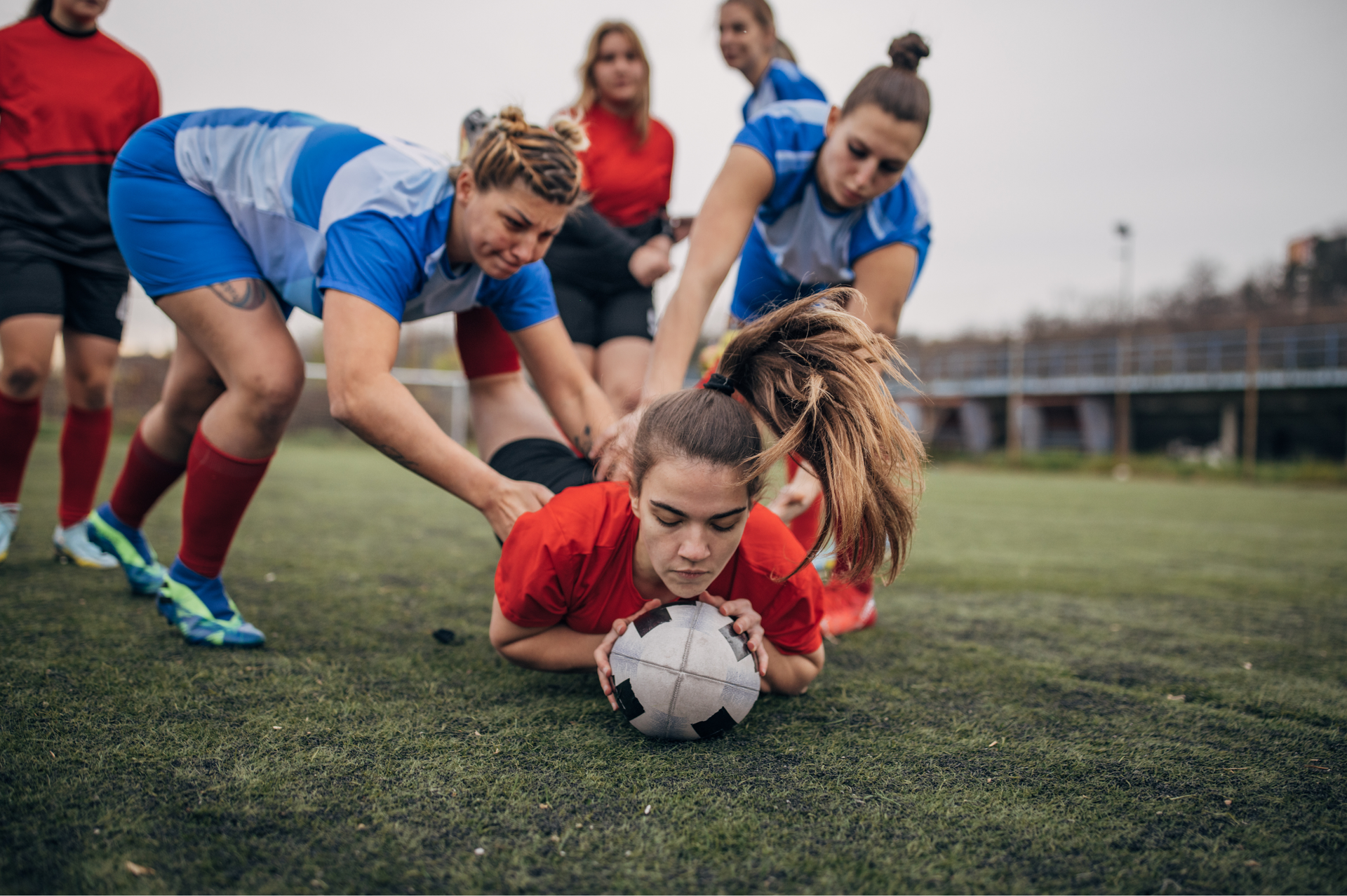 Women's rugby players in blue and red jerseys huddled around a ball on a green field.