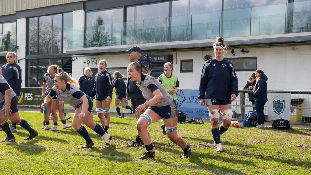 Women's rugby team training outdoors. Players in navy and gray uniforms, focused and athletic, on a grassy field.