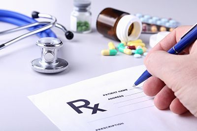 Doctor's hand writing a prescription, with a stethoscope, pills, and medicine bottles on a desk.
