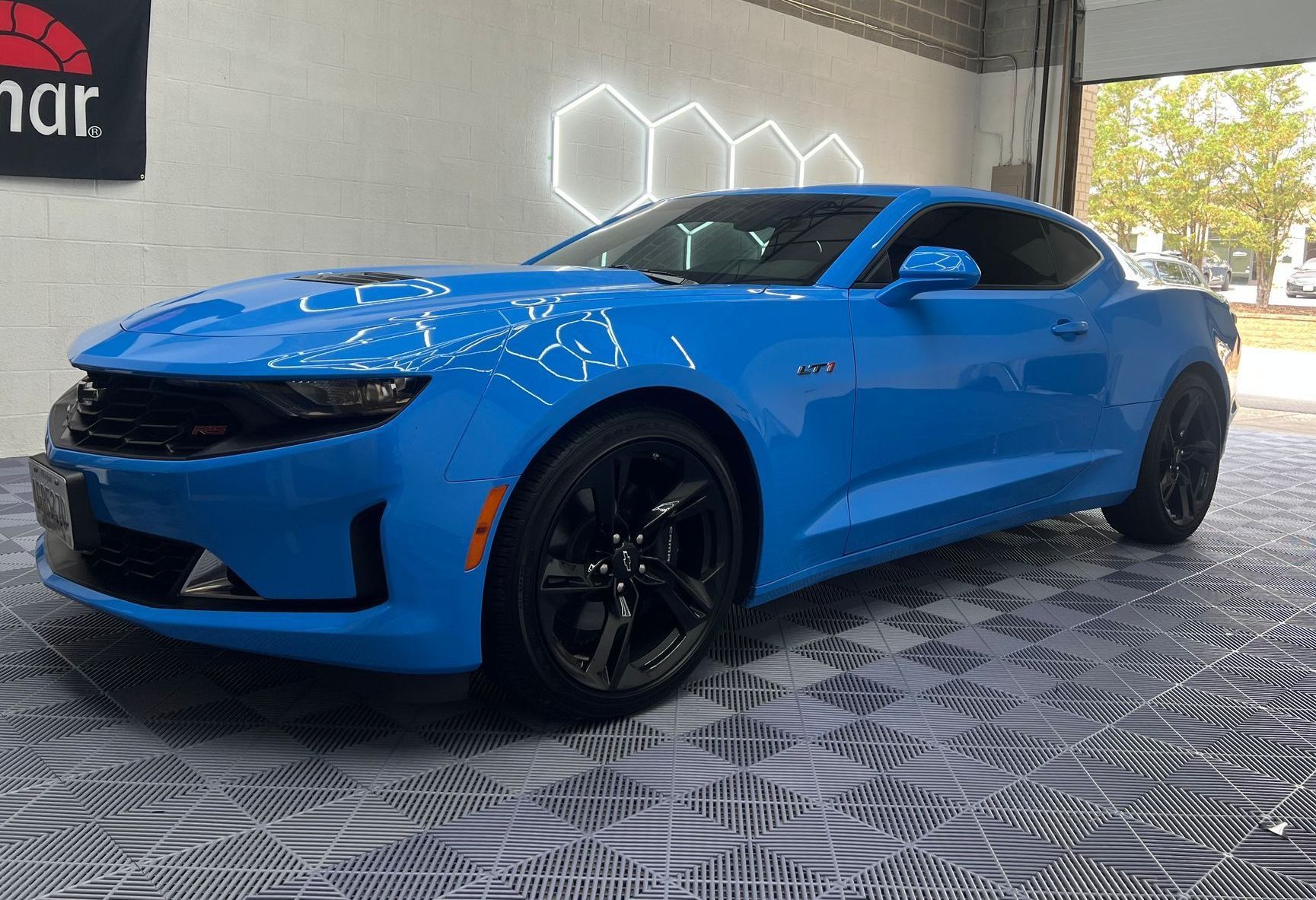 Bright blue Chevrolet Camaro with black wheels, parked in a garage with hexagon-shaped lights, against a light gray patterned floor.