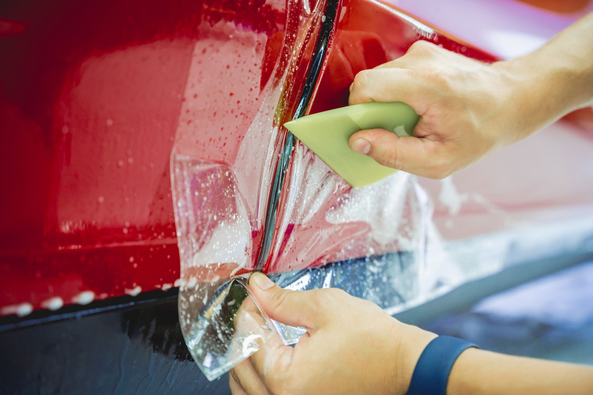 A person is applying a protective film to a red car.