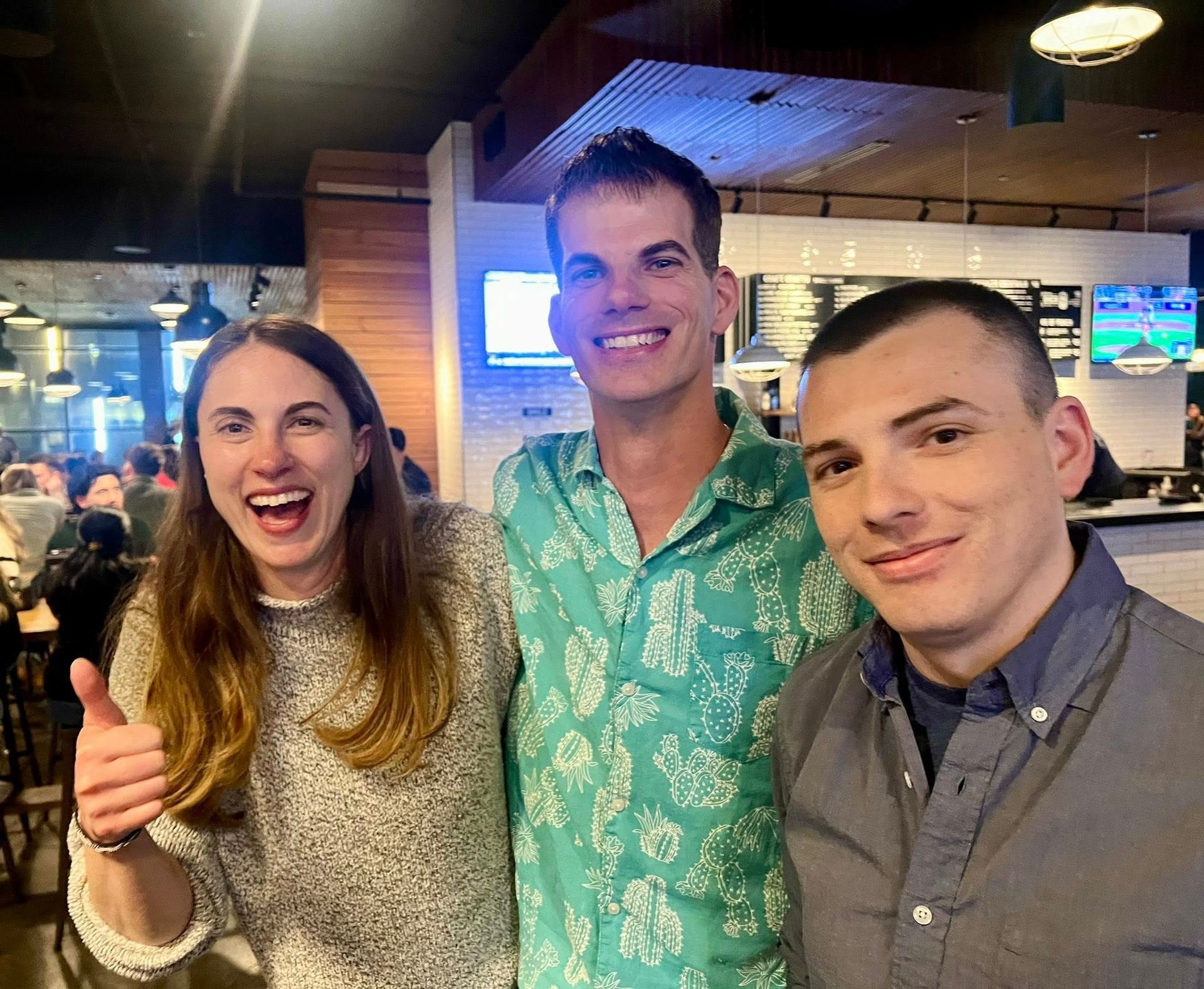 Three people smiling inside a restaurant: a woman gives a thumbs up, a man in a patterned shirt, and a man in a gray shirt.