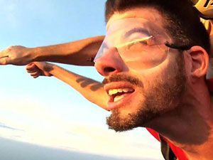 Man in freefall, wearing goggles, arms outstretched, smiling, blue sky background.