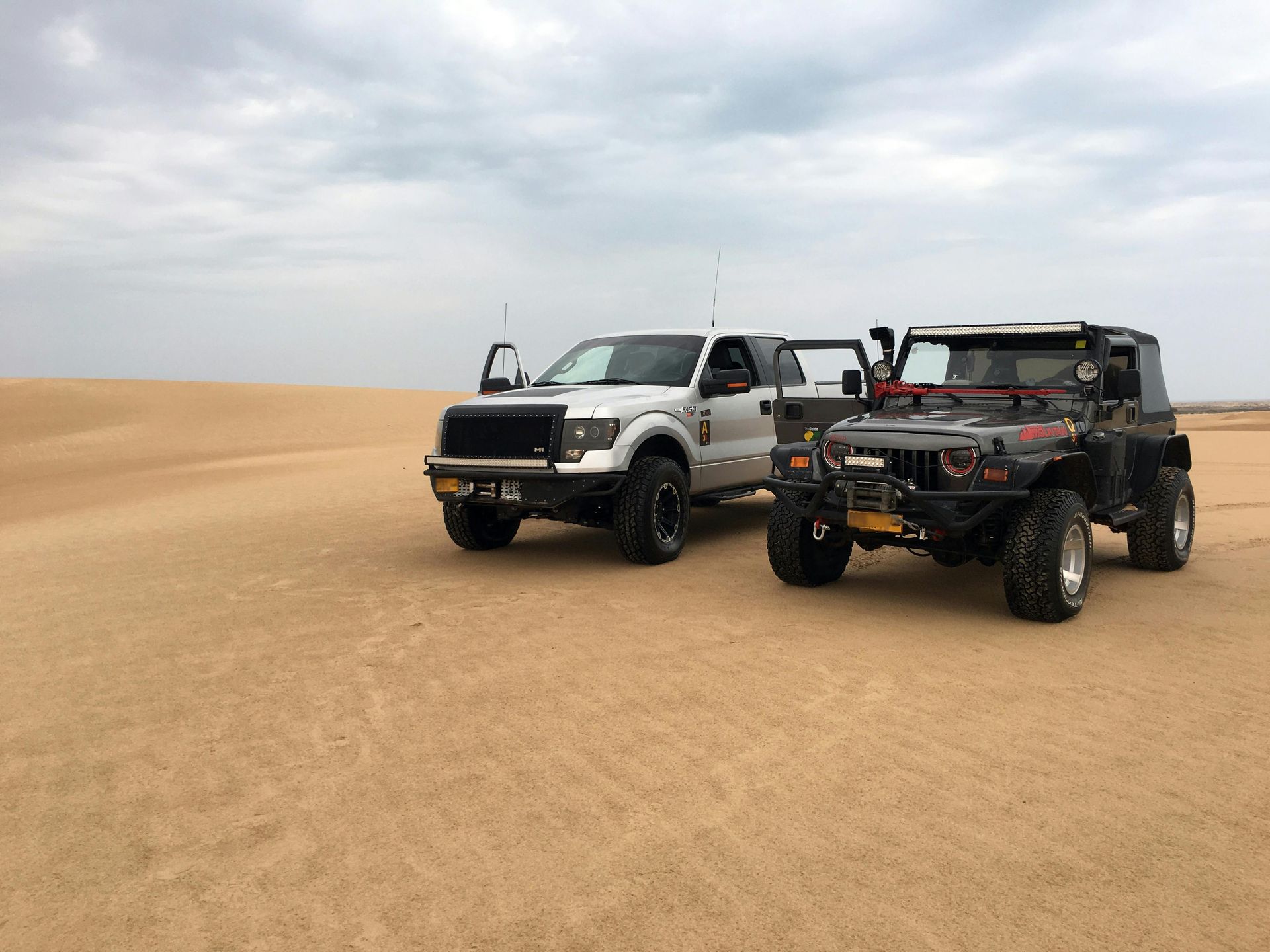 Silver pickup truck and black Jeep parked on a sand dune under a cloudy sky.