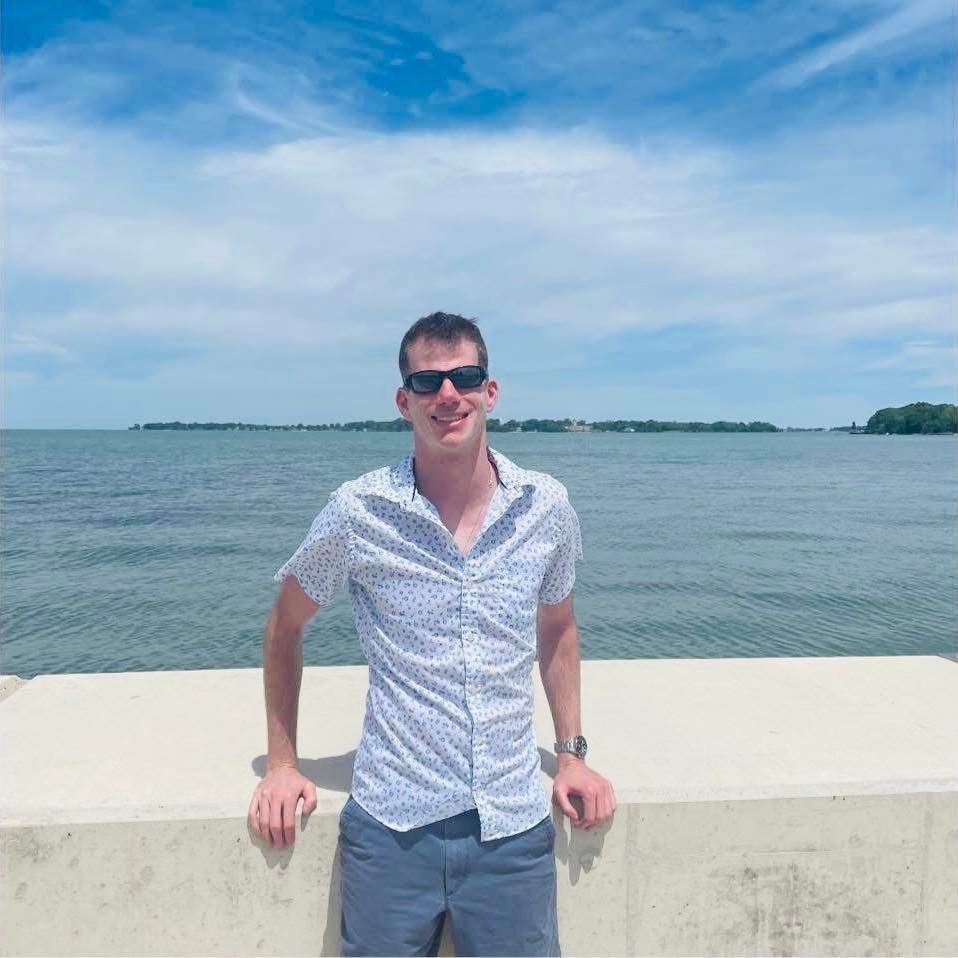 Man in sunglasses and patterned shirt, leaning on concrete wall overlooking water under a blue sky.