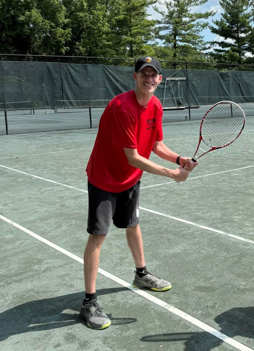 Man in red shirt and black shorts playing tennis on a green court, holding a racket.