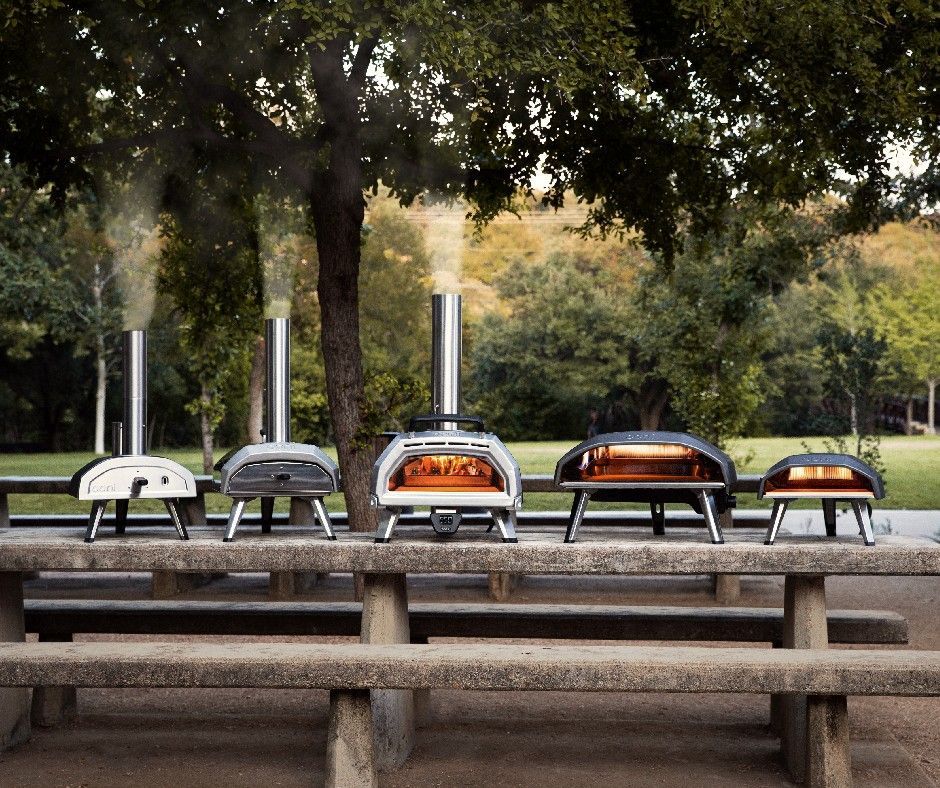 Uma fileira de fornos de pizza está alinhada em uma mesa de piquenique.