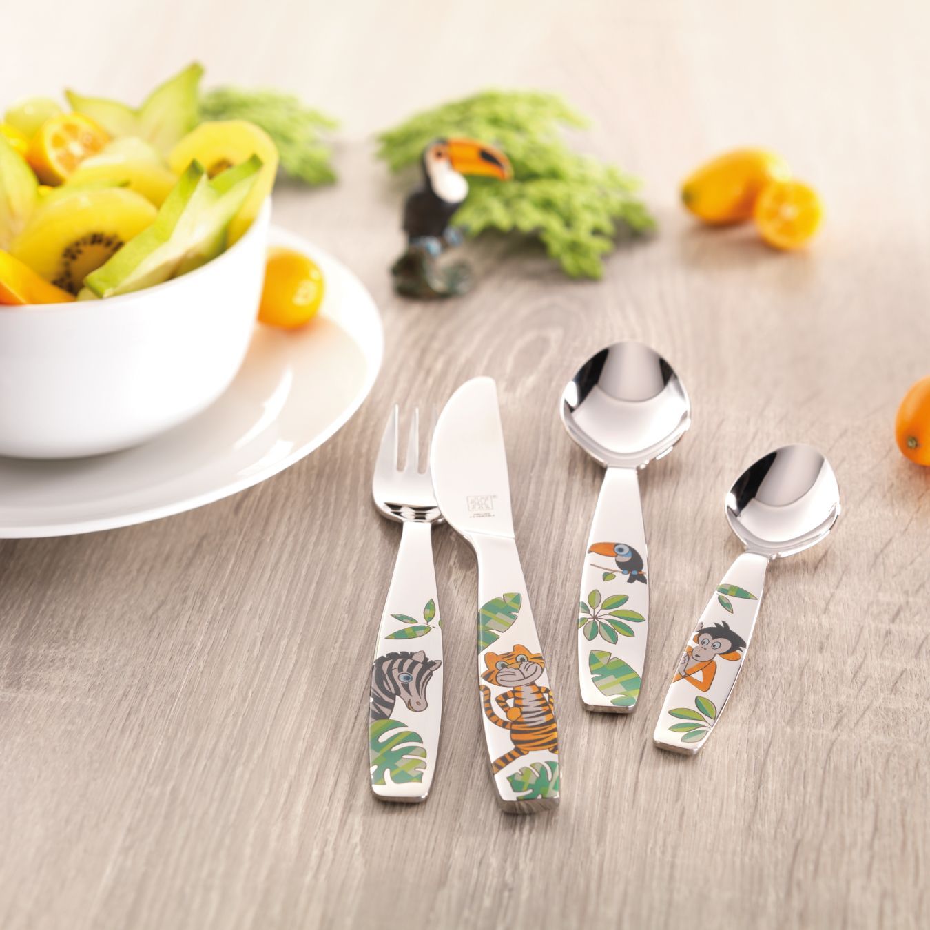 Talheres infantis com animais nas alças e uma tigela de frutas ao fundo