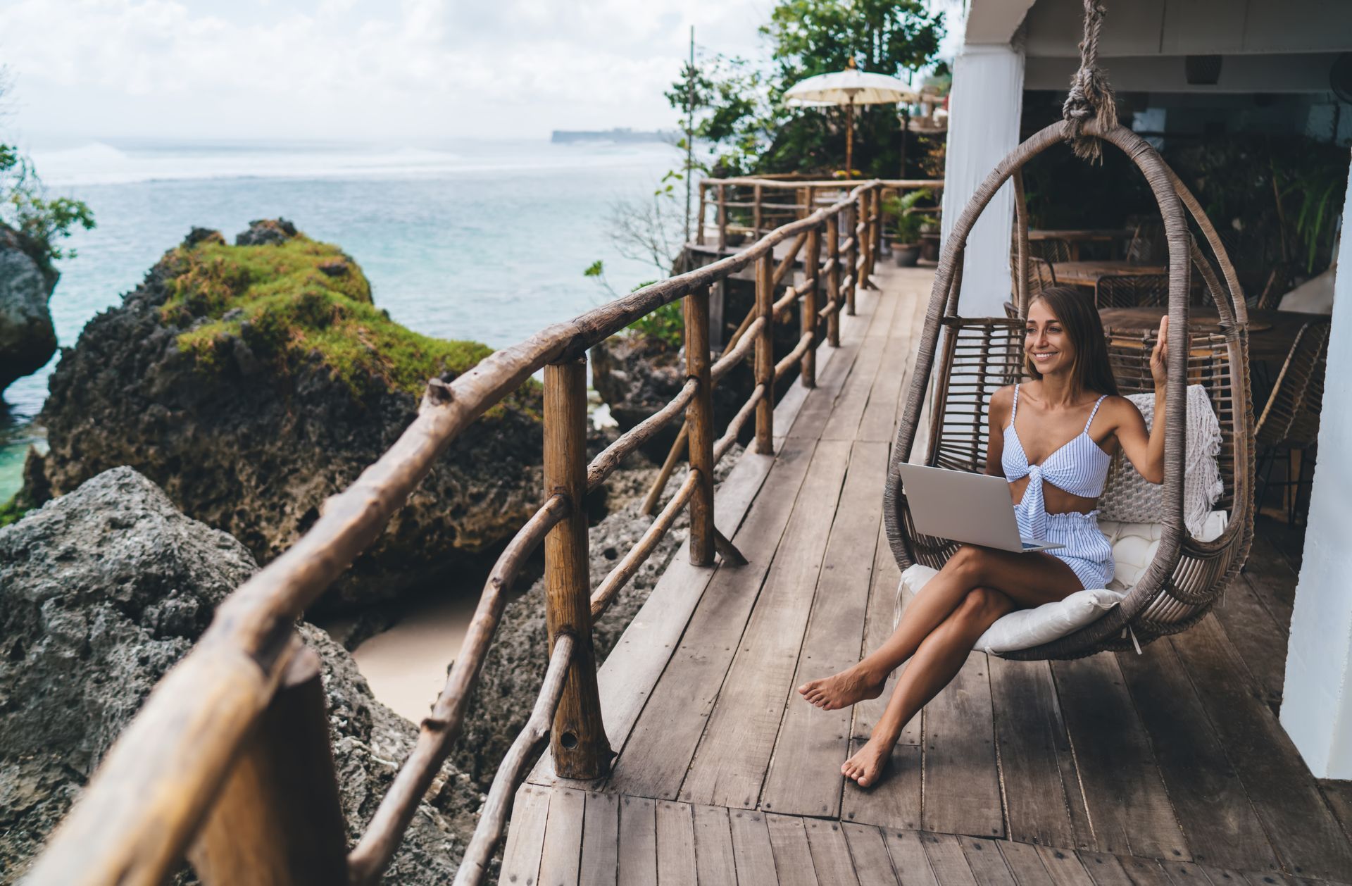 A woman is sitting in a hanging chair on a balcony with a laptop.