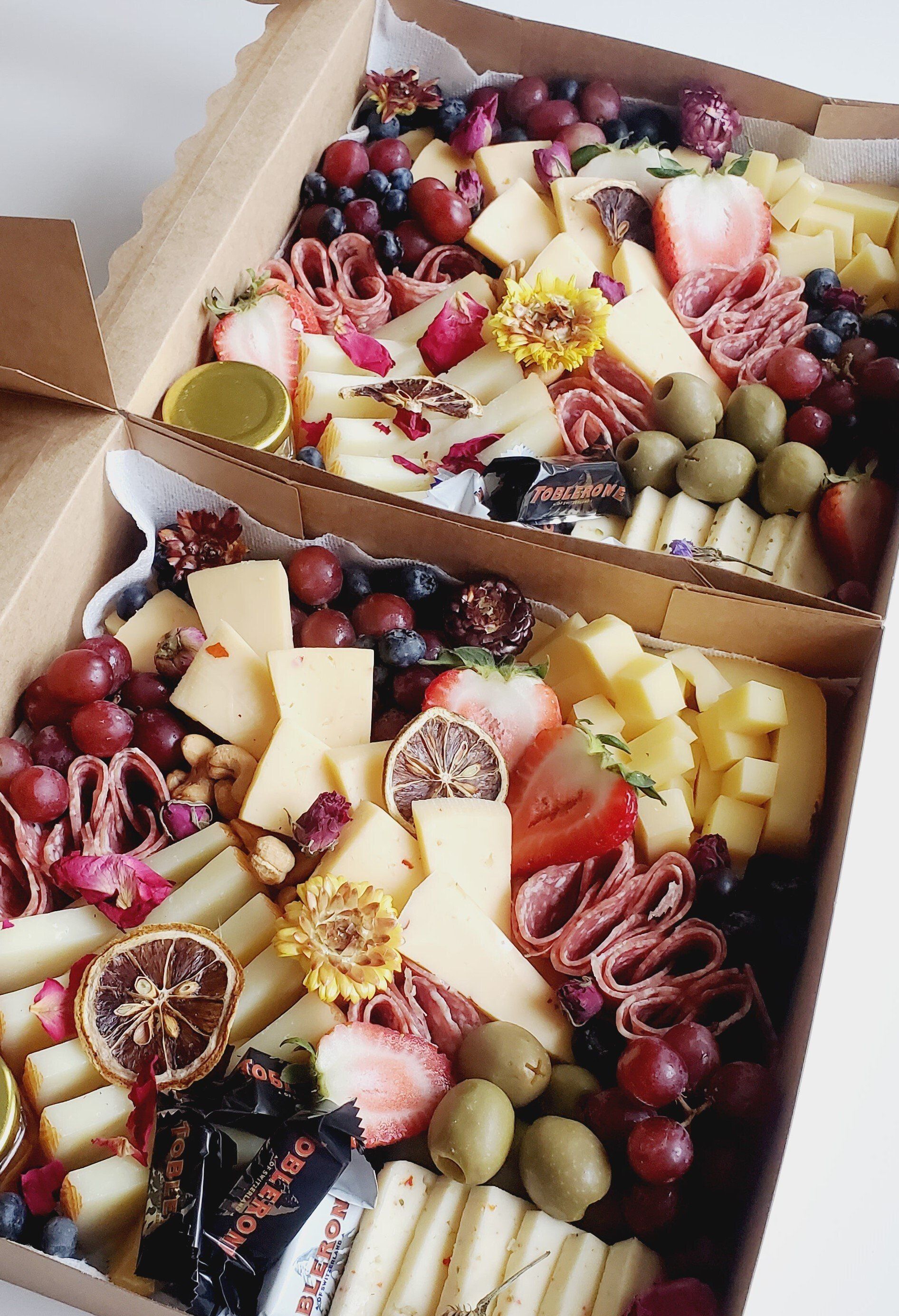 Two boxes filled with cheese , grapes , olives and strawberries.