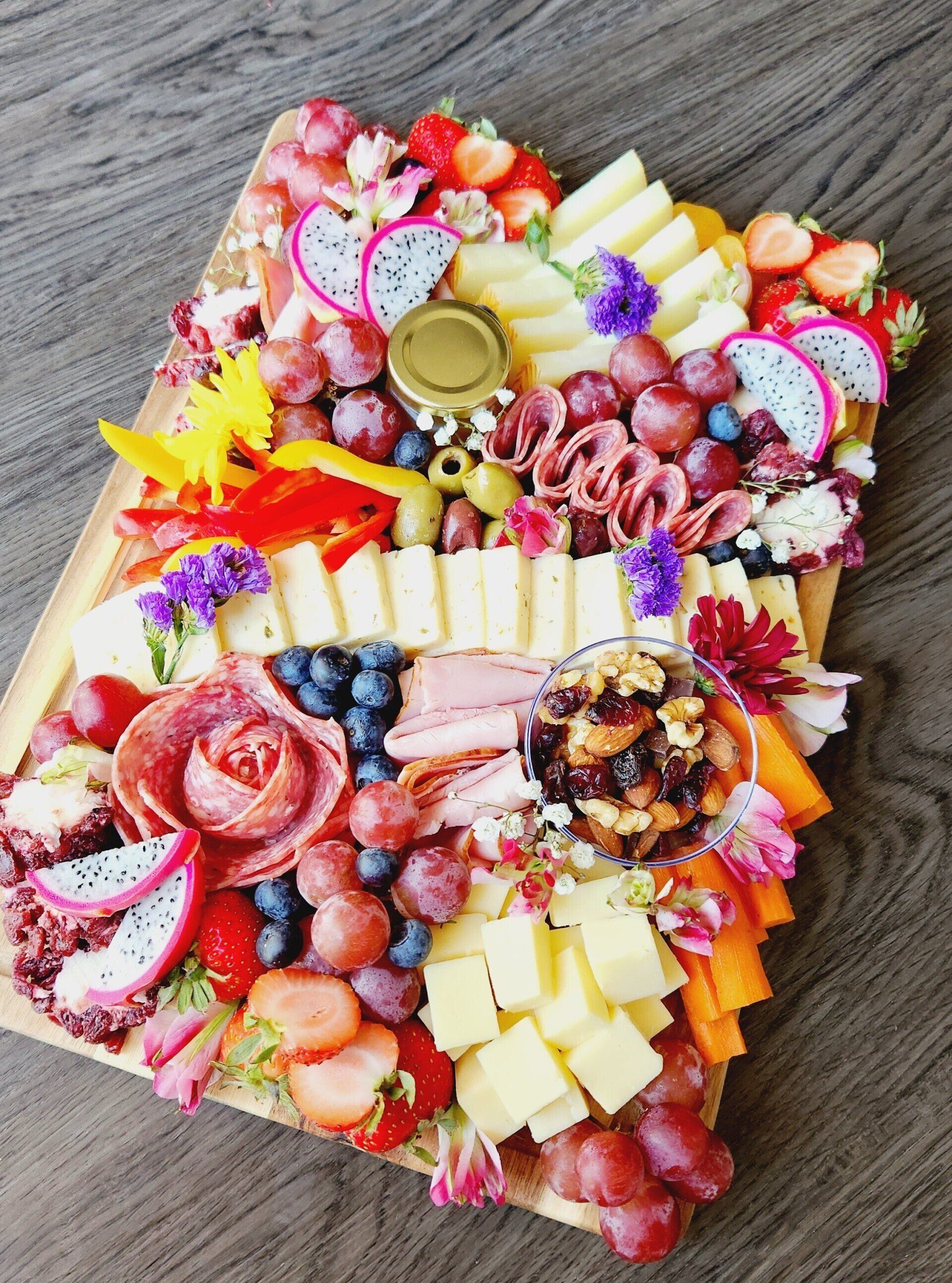 A cutting board filled with fruits , cheeses , meats and flowers on a wooden table.