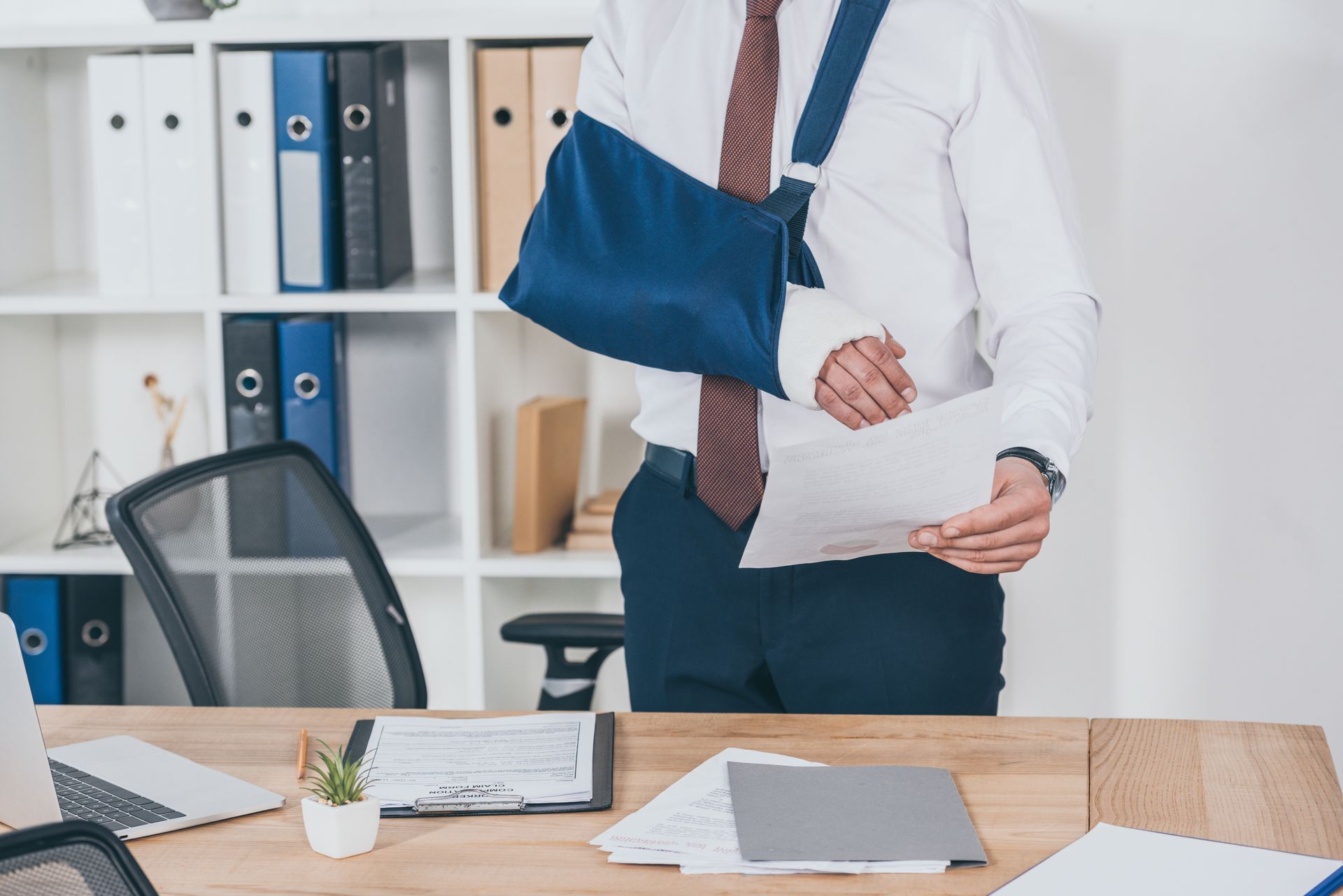 A male worker with a broken arm on a sling stands in front of a table, while holding a document.