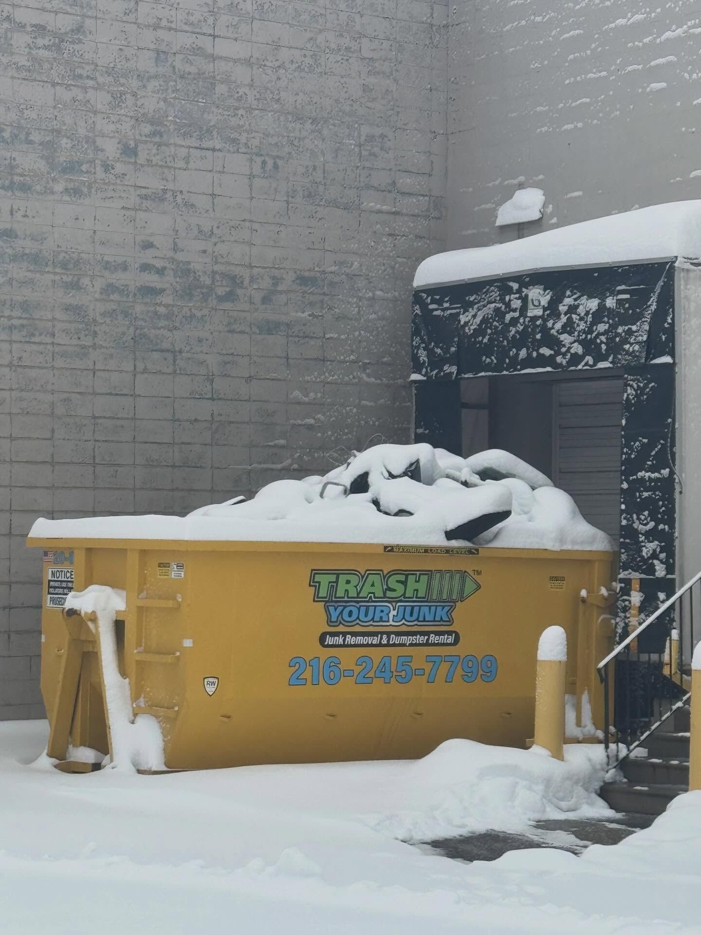 A bright yellow dumpster sits on a driveway in front of a brick house during a renovation project.