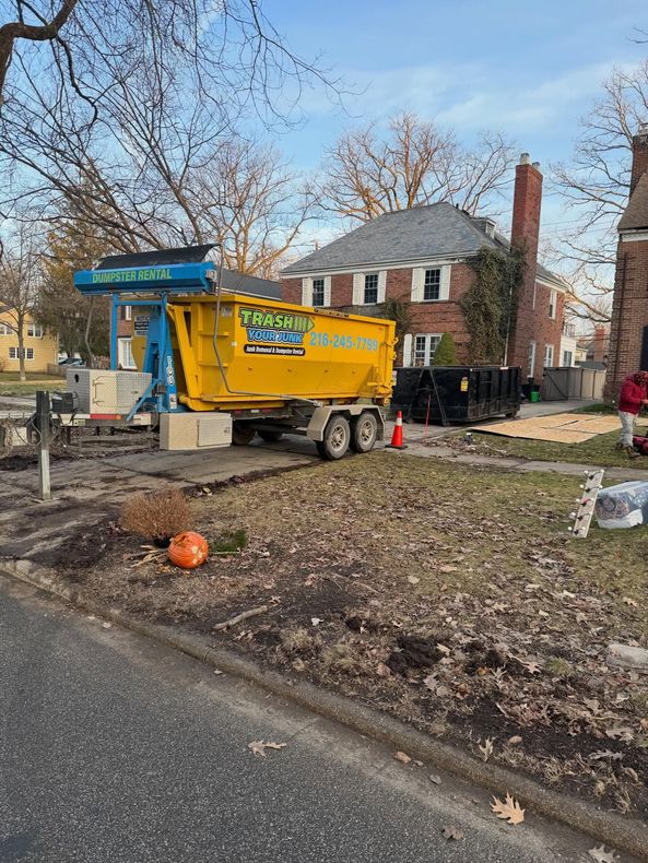 A bright yellow dumpster sits on a driveway in front of a brick house during a renovation project.